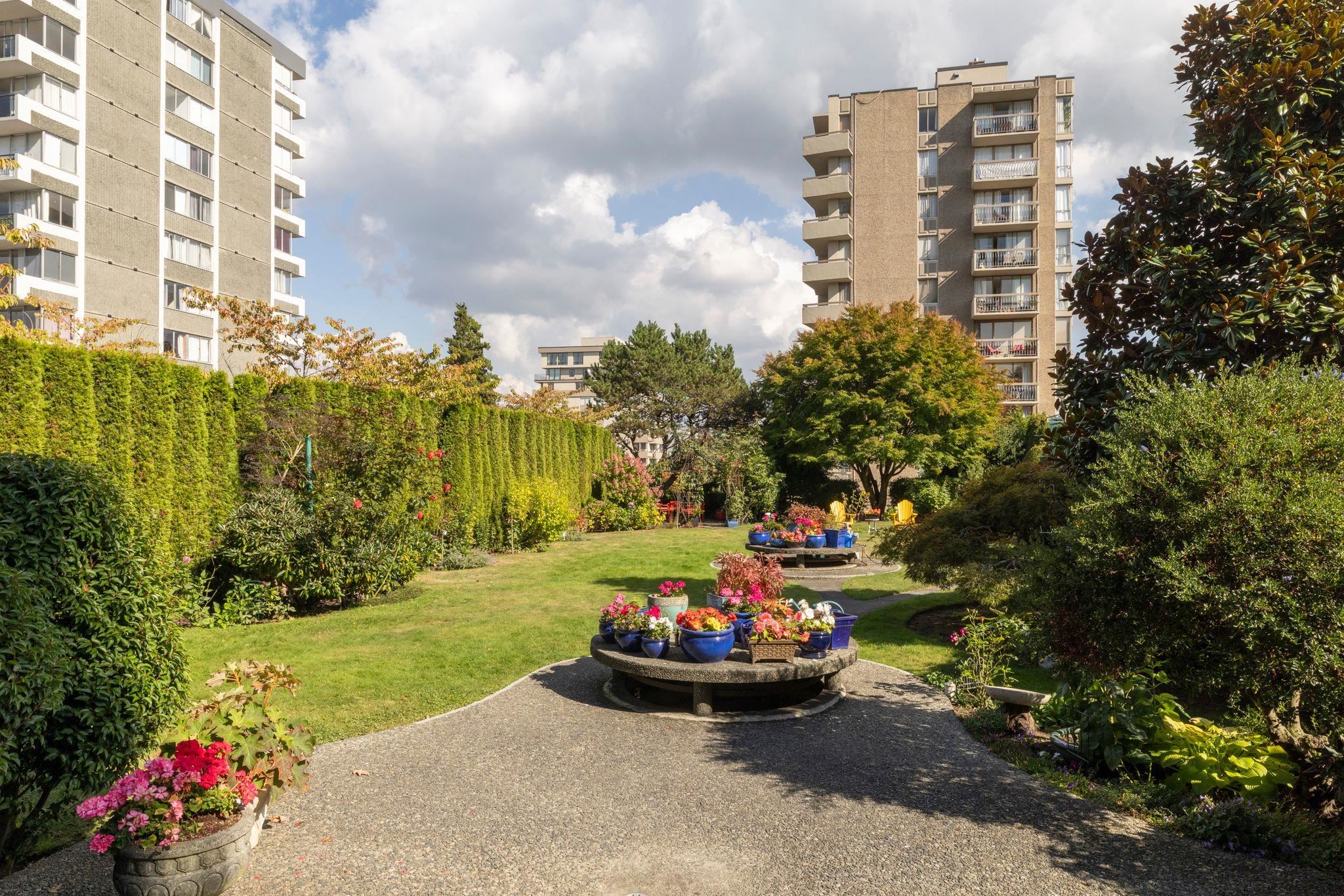 Apartment courtyard garden with a lawn, flowering planters, and paved paths between residential towers.