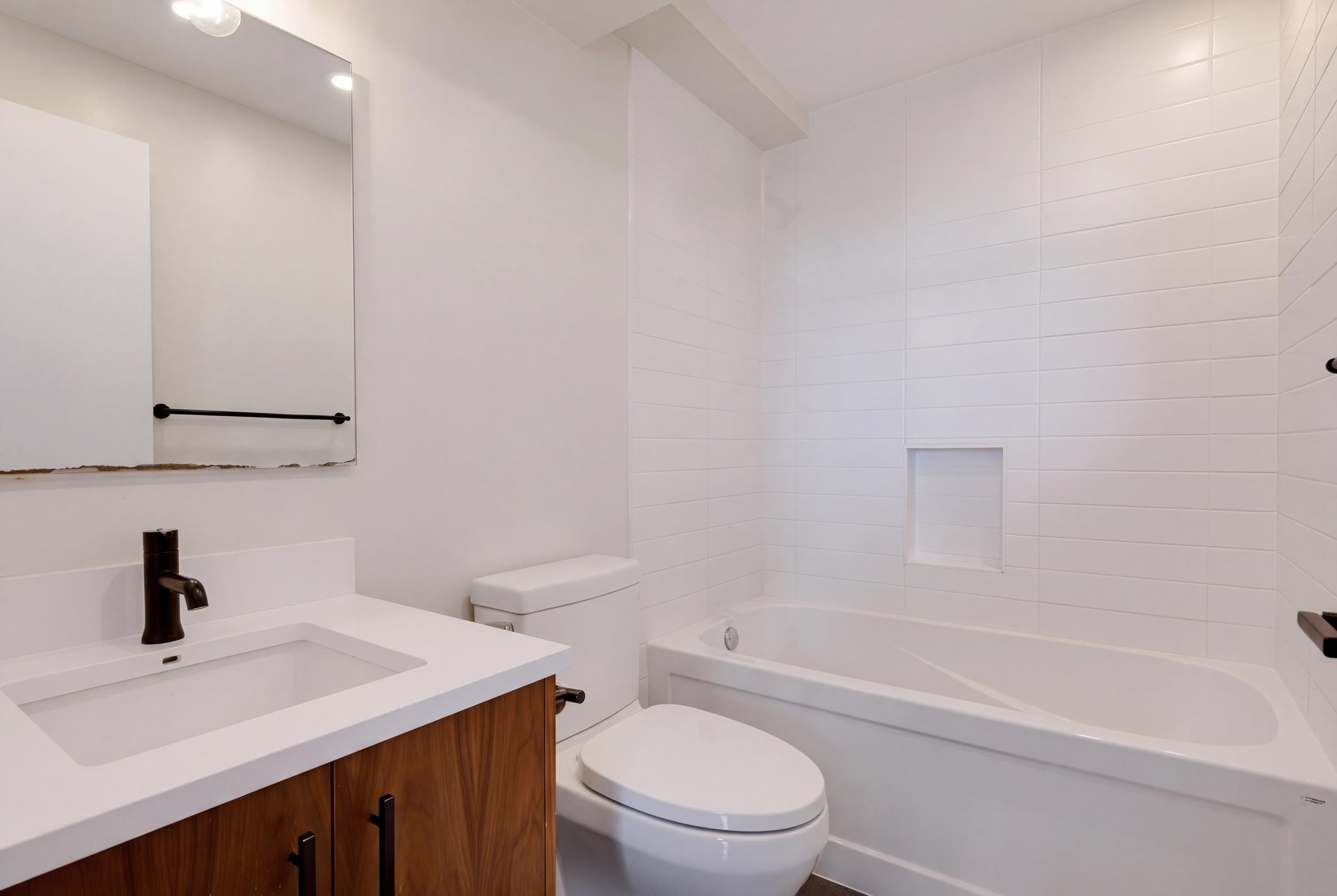 Modern bathroom with a white tiled bathtub, recessed niche, and wood vanity with dark faucet.