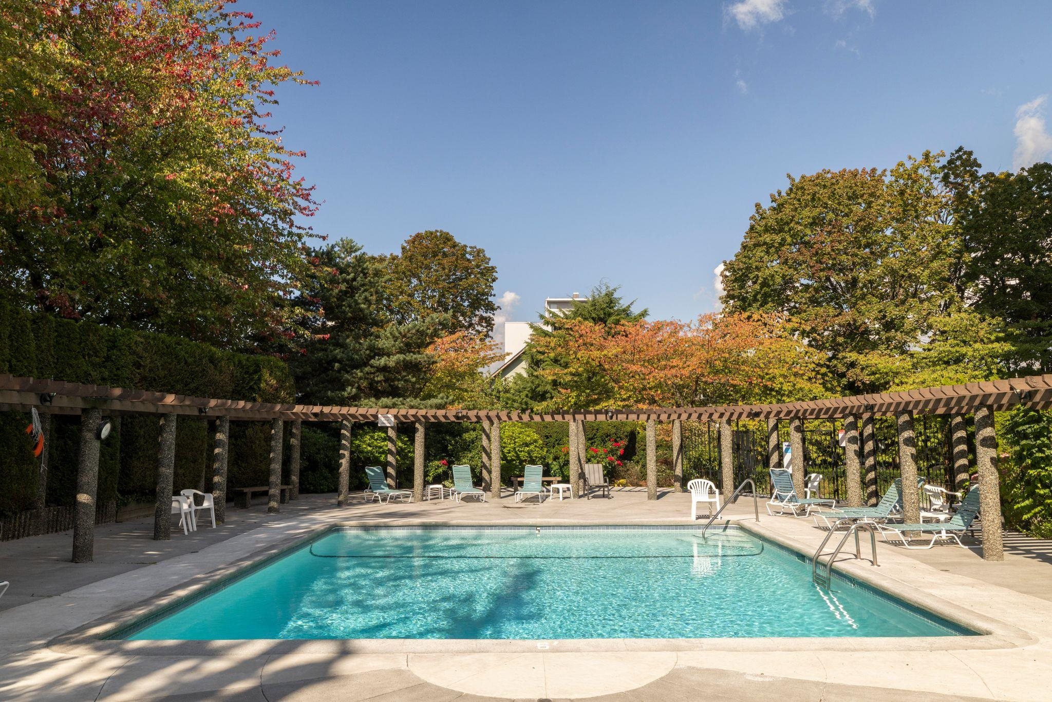 Outdoor pool beneath a wooden pergola, surrounded by lounge chairs and autumn trees.