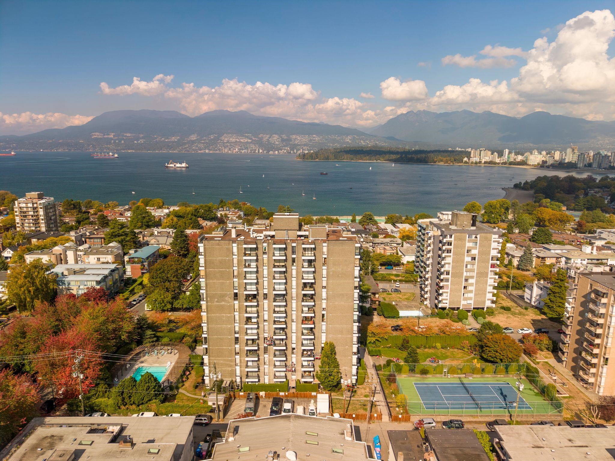 Aerial view of a Vancouver condominium complex with a tennis court, pool, waterfront and mountain backdrop.