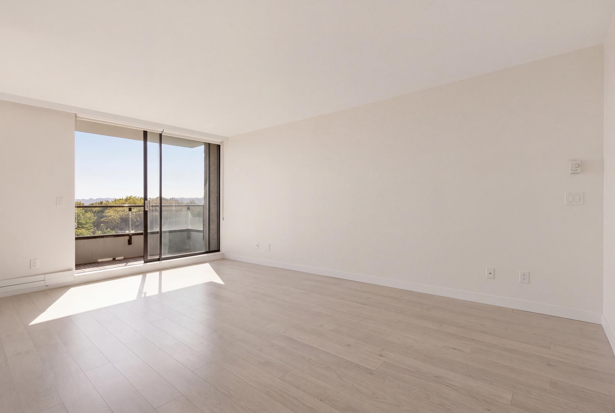 Empty living room with pale plank flooring and sunny sliding doors opening onto a balcony.