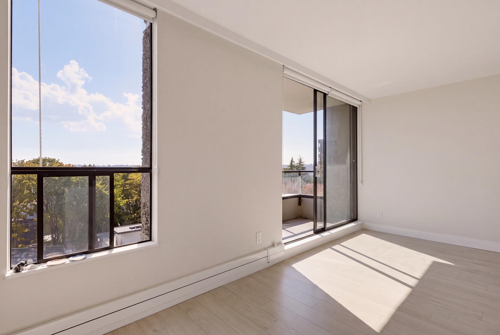 Vacant sunlit room with pale wood floors, a large window, and a sliding door to the balcony.
