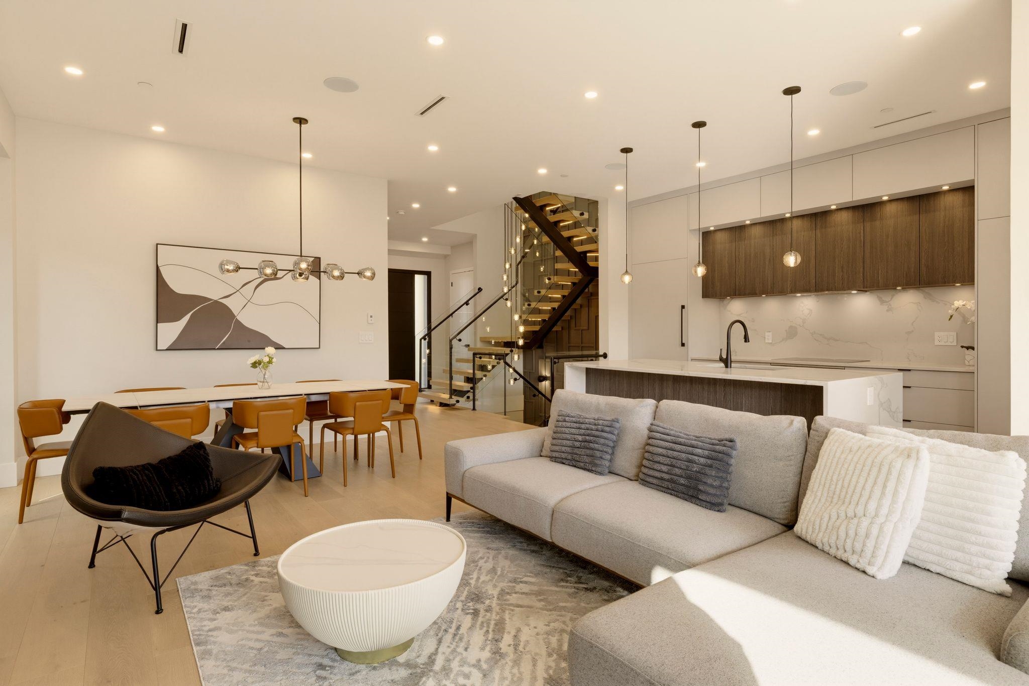 Open-plan living room with a gray sectional, dining area, kitchen island and illuminated glass staircase.