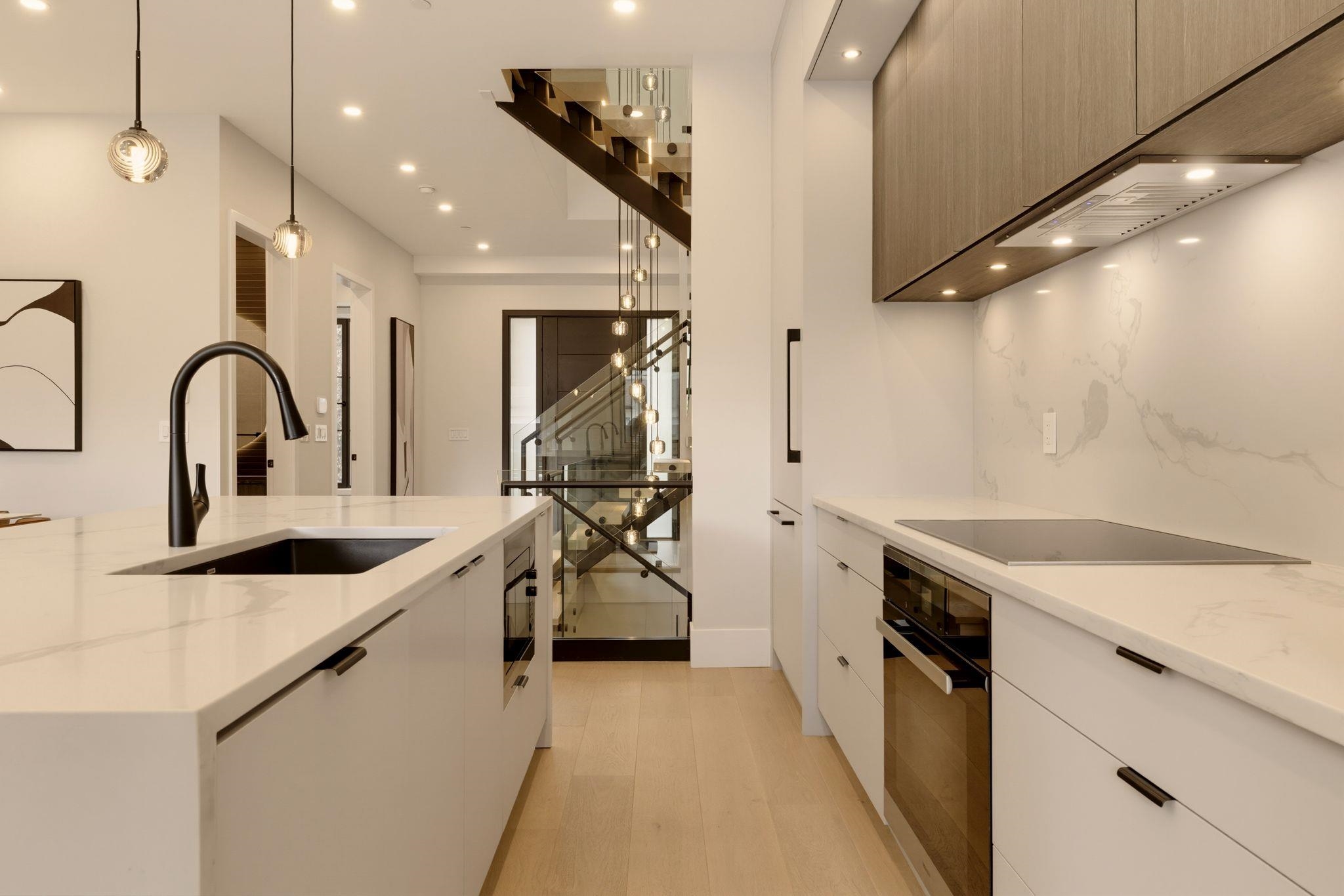 Modern kitchen with marble-look counters, dark wood cabinets, and a glass-railed staircase beyond.
