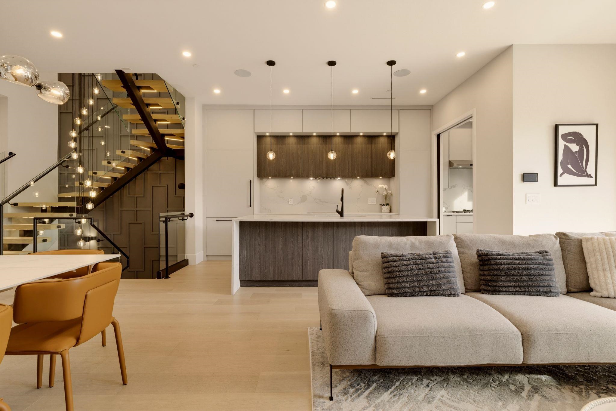Open-plan kitchen and living area with a large island, sectional sofa, and glass-railed staircase.