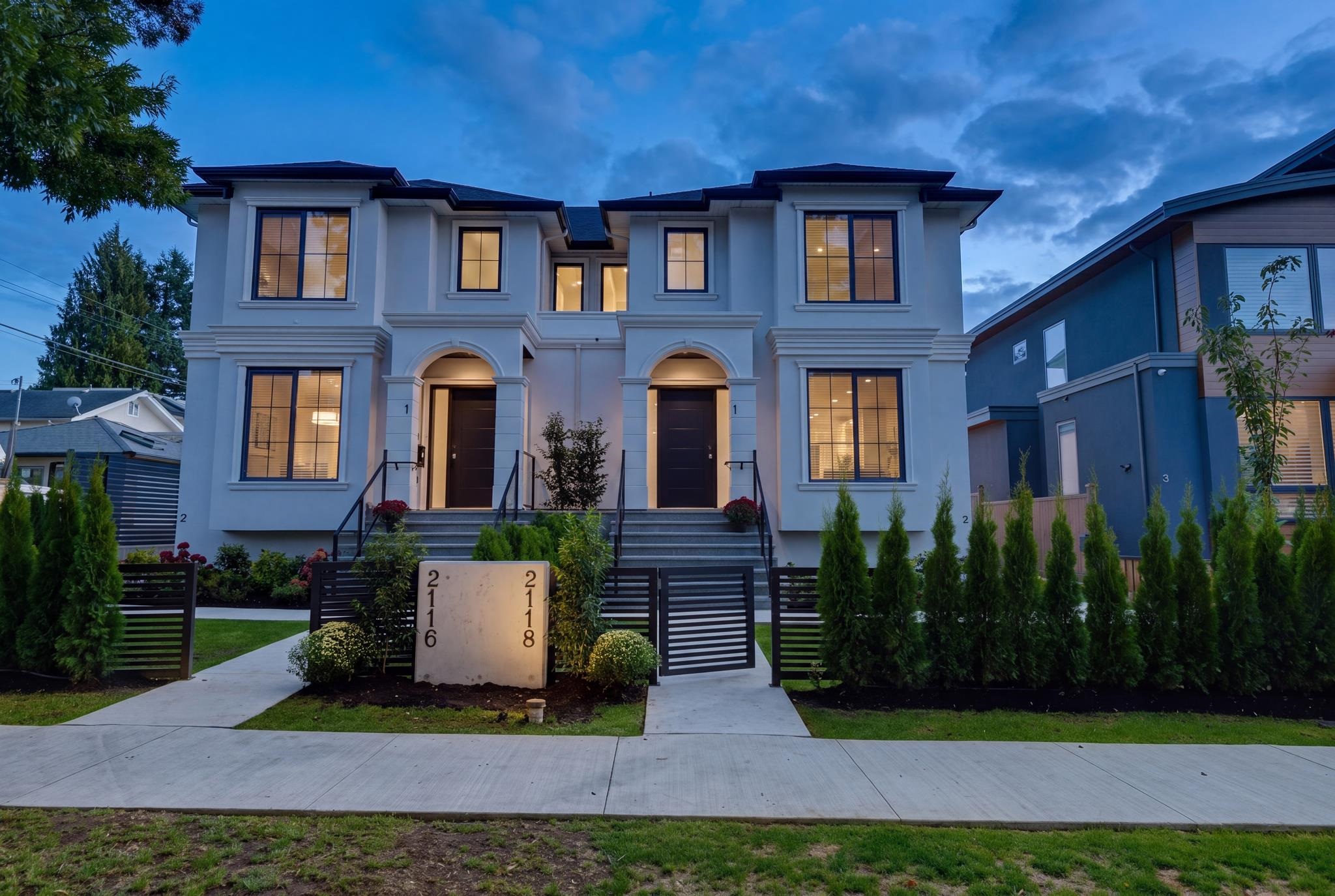 White two-storey duplex at dusk, with twin arched entrances and illuminated windows.