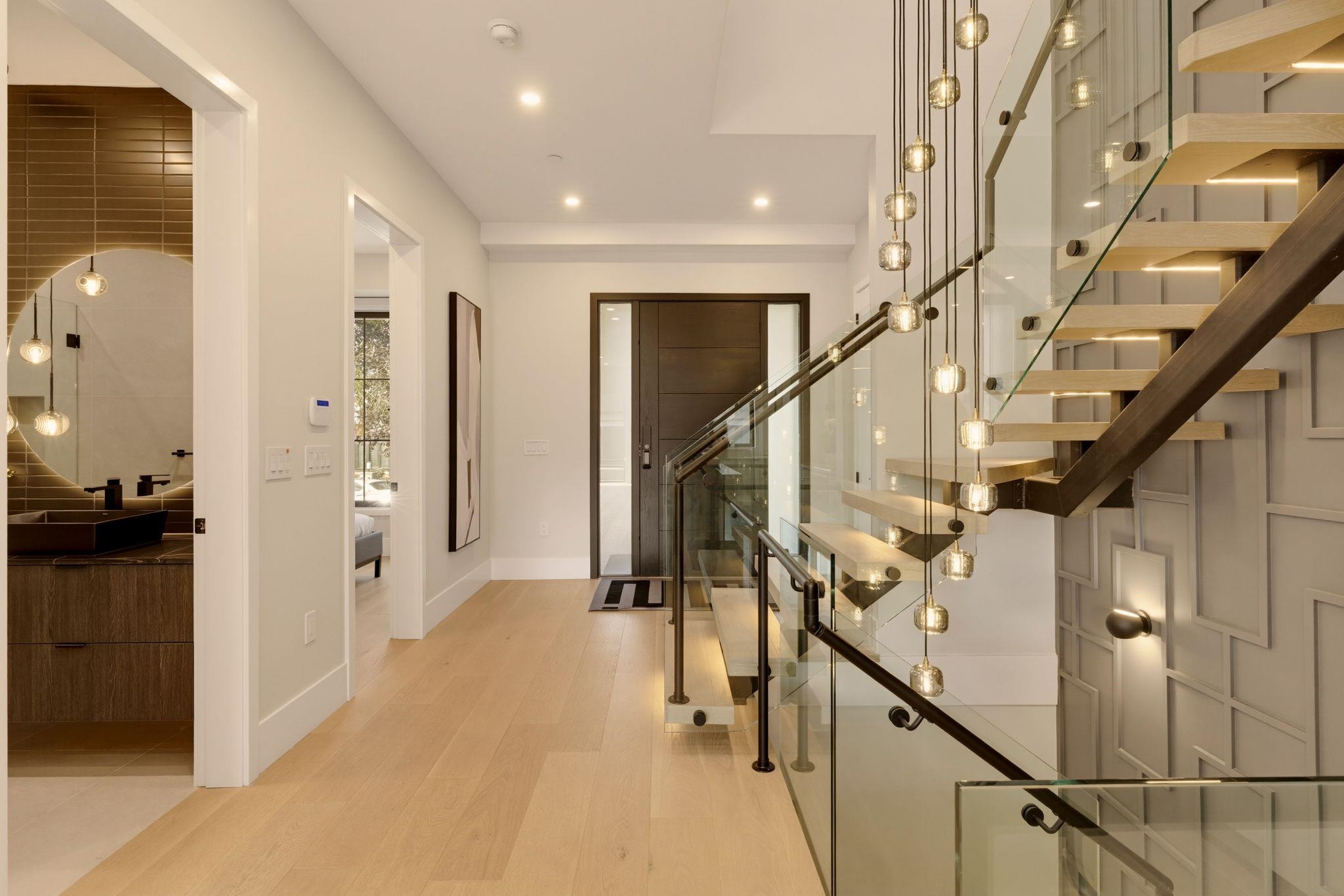 Entry hall with a glass-railed floating staircase and pendant lights beside a dark-tiled powder room.