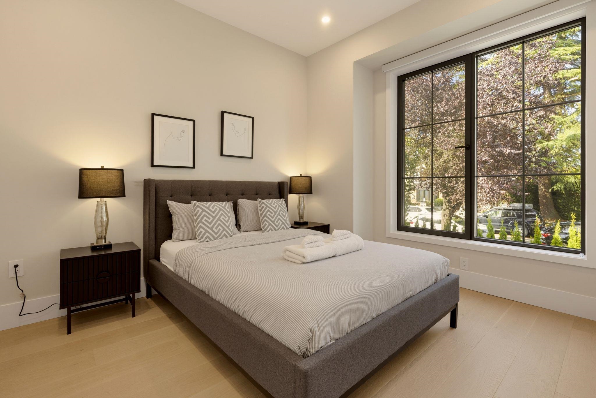 Staged bedroom with an upholstered bed and a large window overlooking leafy trees.