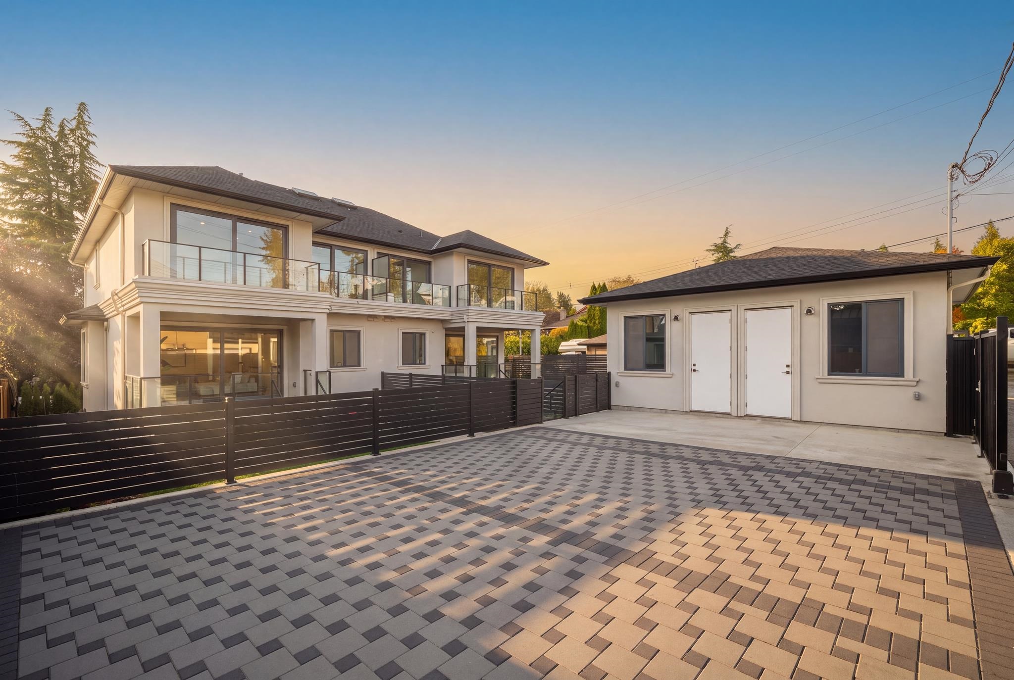Modern two-story home with glass balcony and detached outbuilding beyond a paved courtyard.