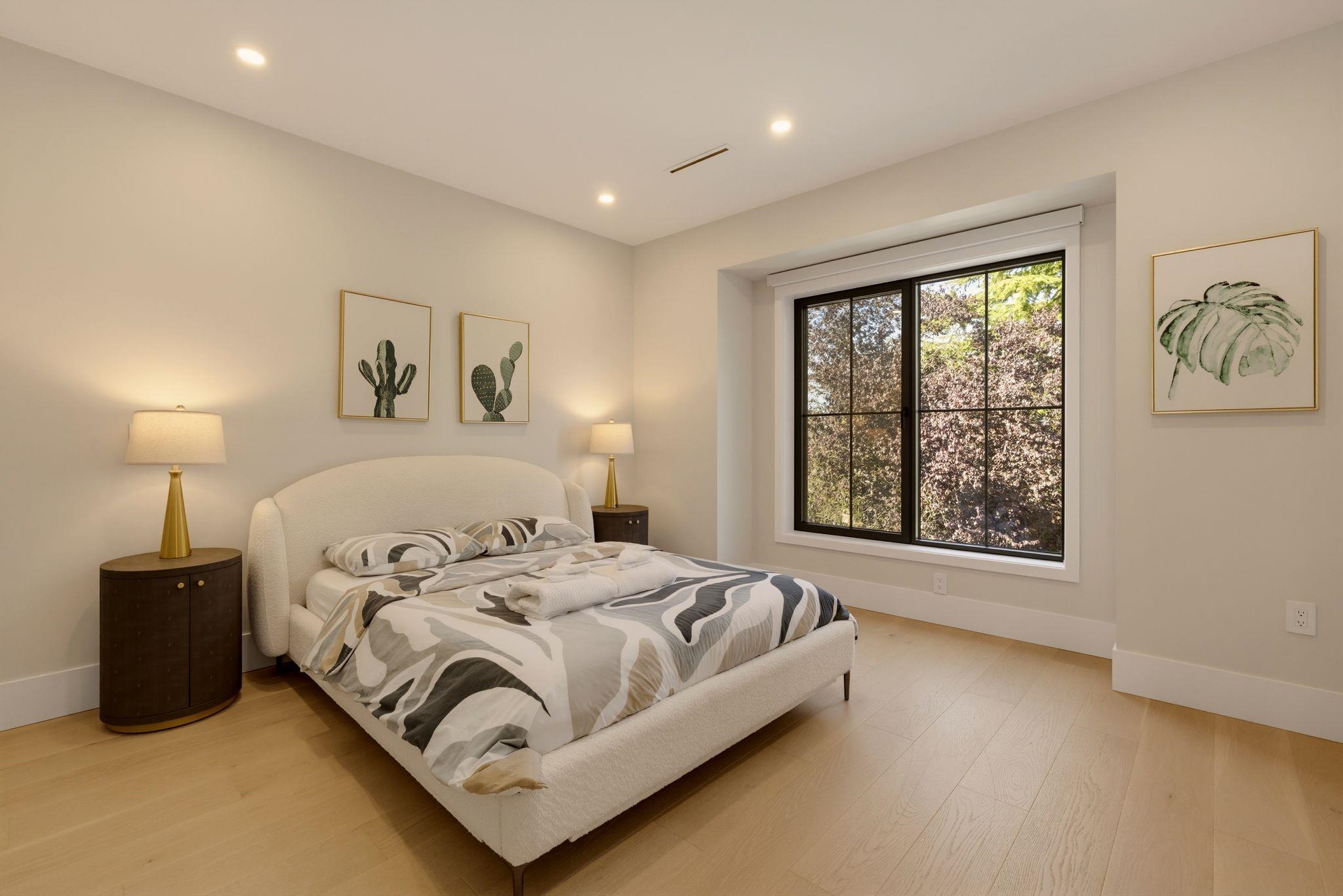 Staged bedroom with a cream bed, patterned bedding, and a large window overlooking leafy trees.