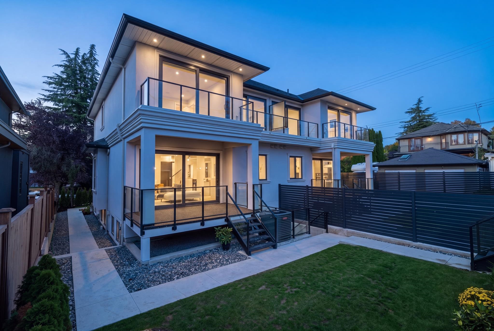 Modern two-story home at dusk with glass-railed balconies, a raised patio, and a fenced lawn.