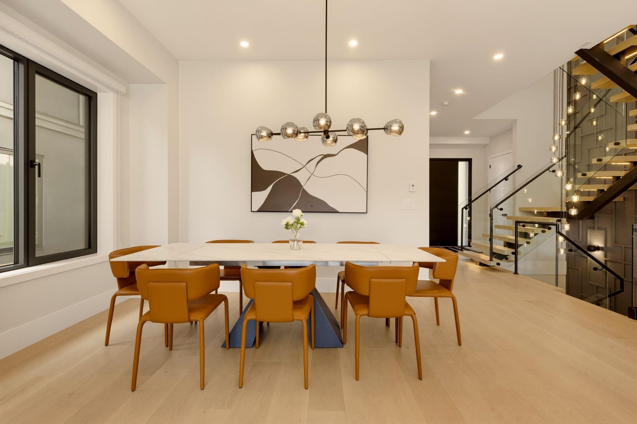Modern dining room with a white table, cognac leather chairs, and glass-railed staircase.