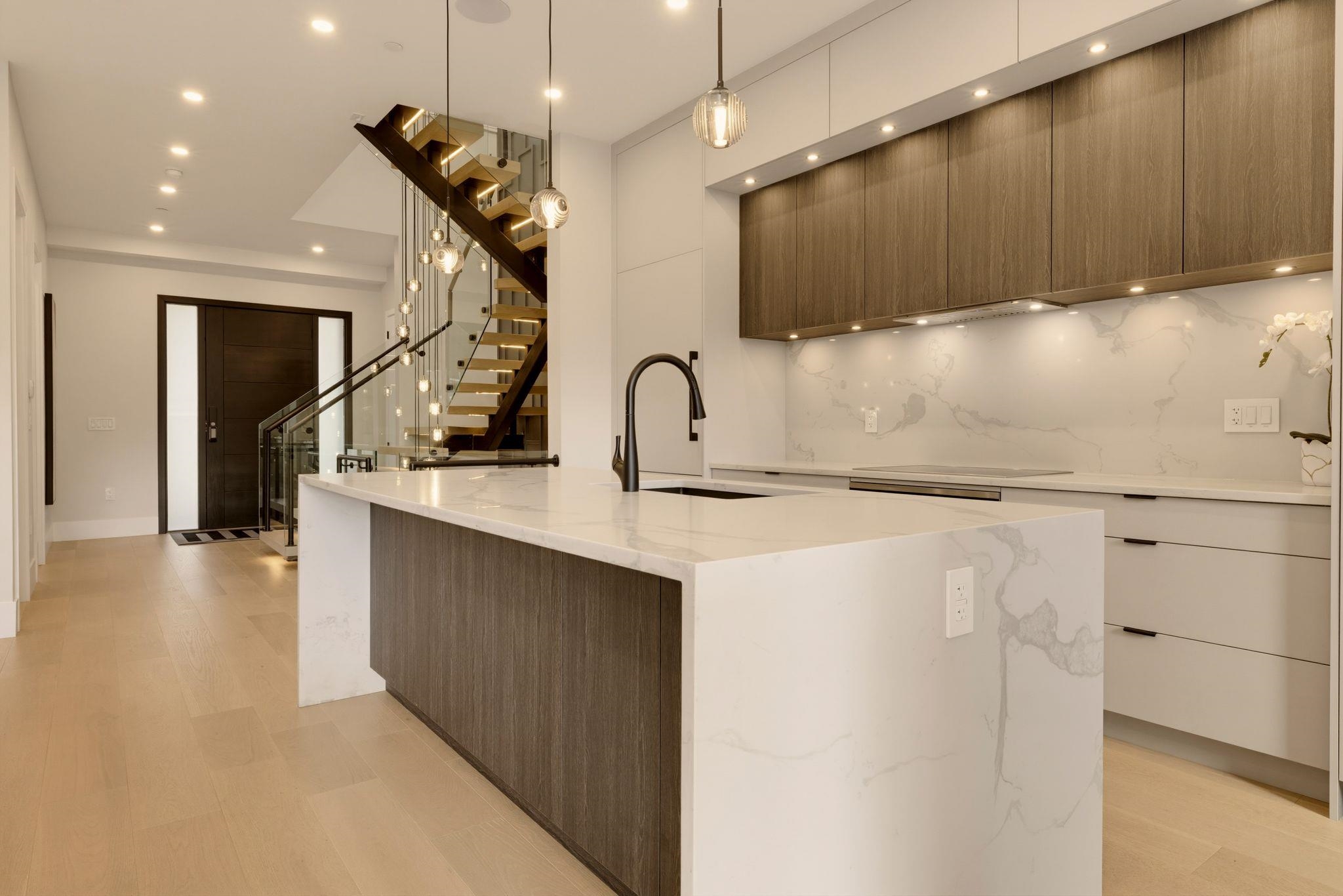 Modern kitchen with a large veined-stone island, dark wood cabinets, and a glass-railed staircase.