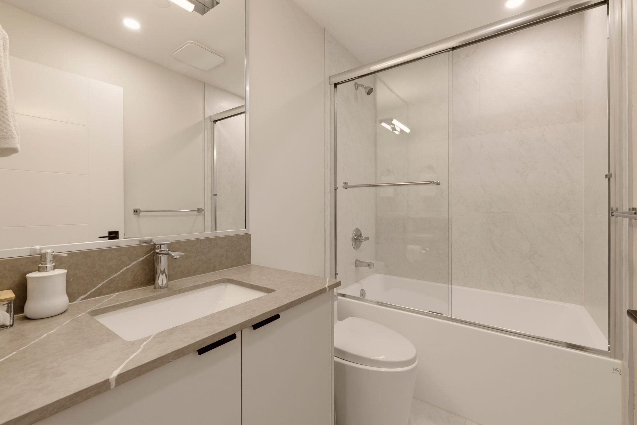 Modern bathroom with a stone vanity, large mirror, and glass-enclosed bathtub.