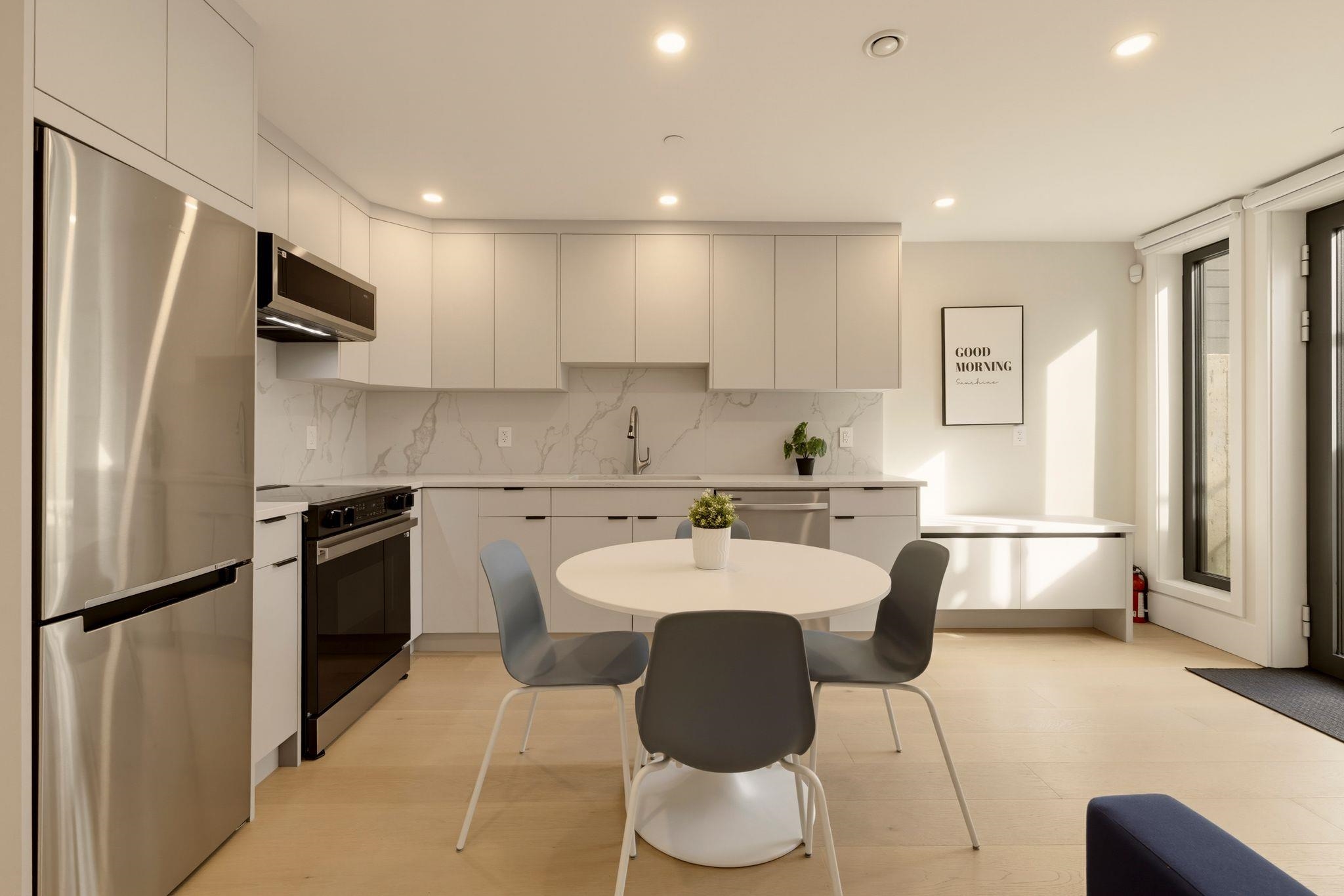 Modern white kitchen with stainless steel appliances, a round dining table, and a sunlit breakfast counter.