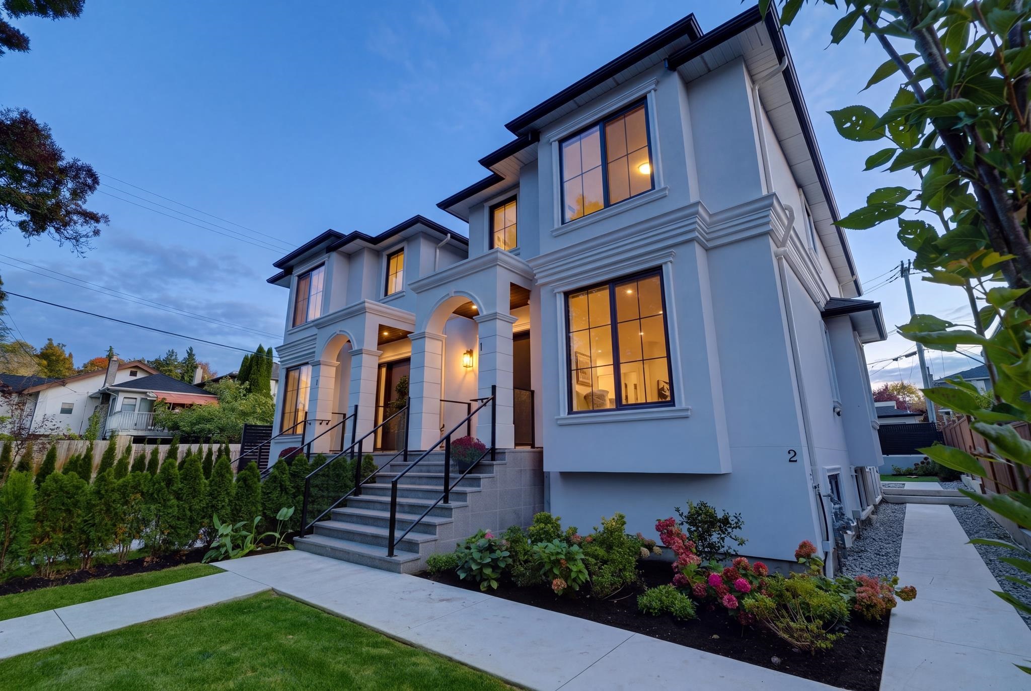 Large white home with arched entry and illuminated windows at dusk, bordered by landscaped flower beds.