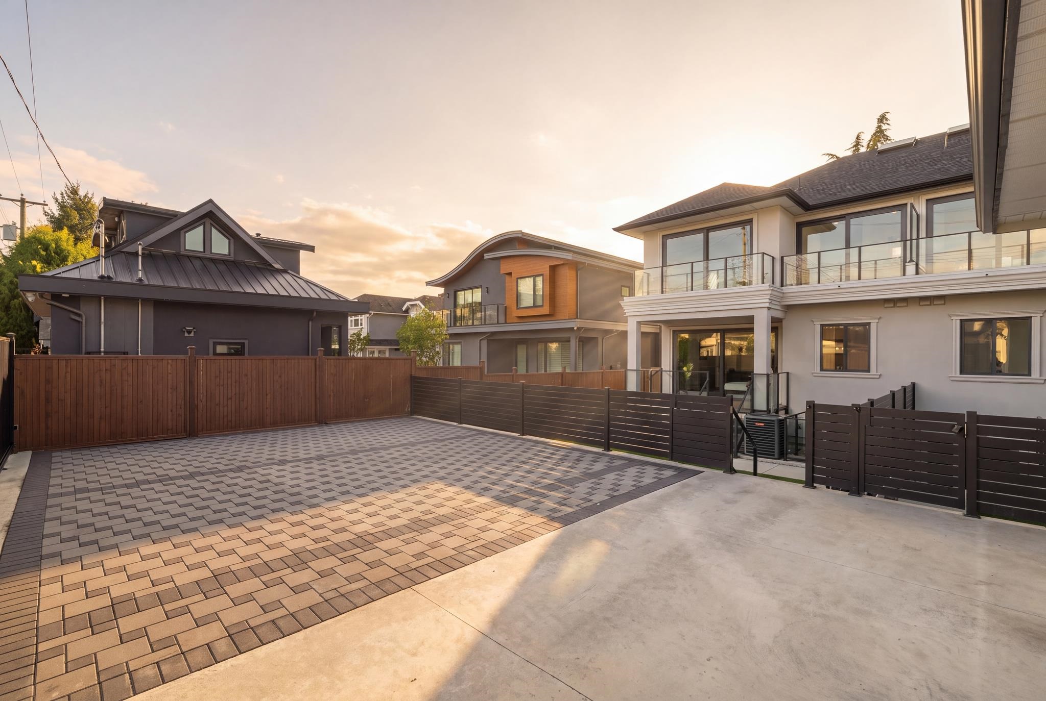 Modern home beside a broad paved patio with privacy fencing and a glass-railed balcony.