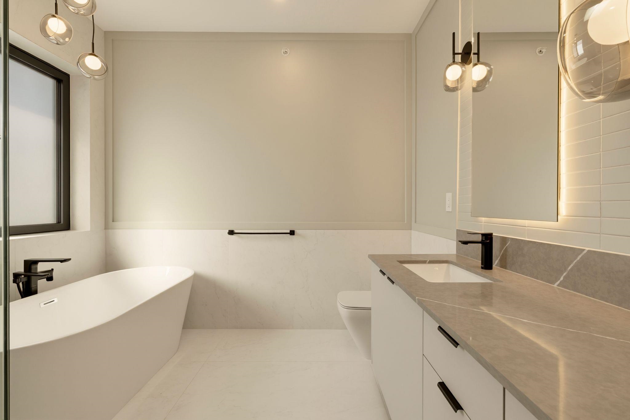 Modern bathroom with a freestanding tub, stone-topped vanity, illuminated mirror, and globe pendant lights.