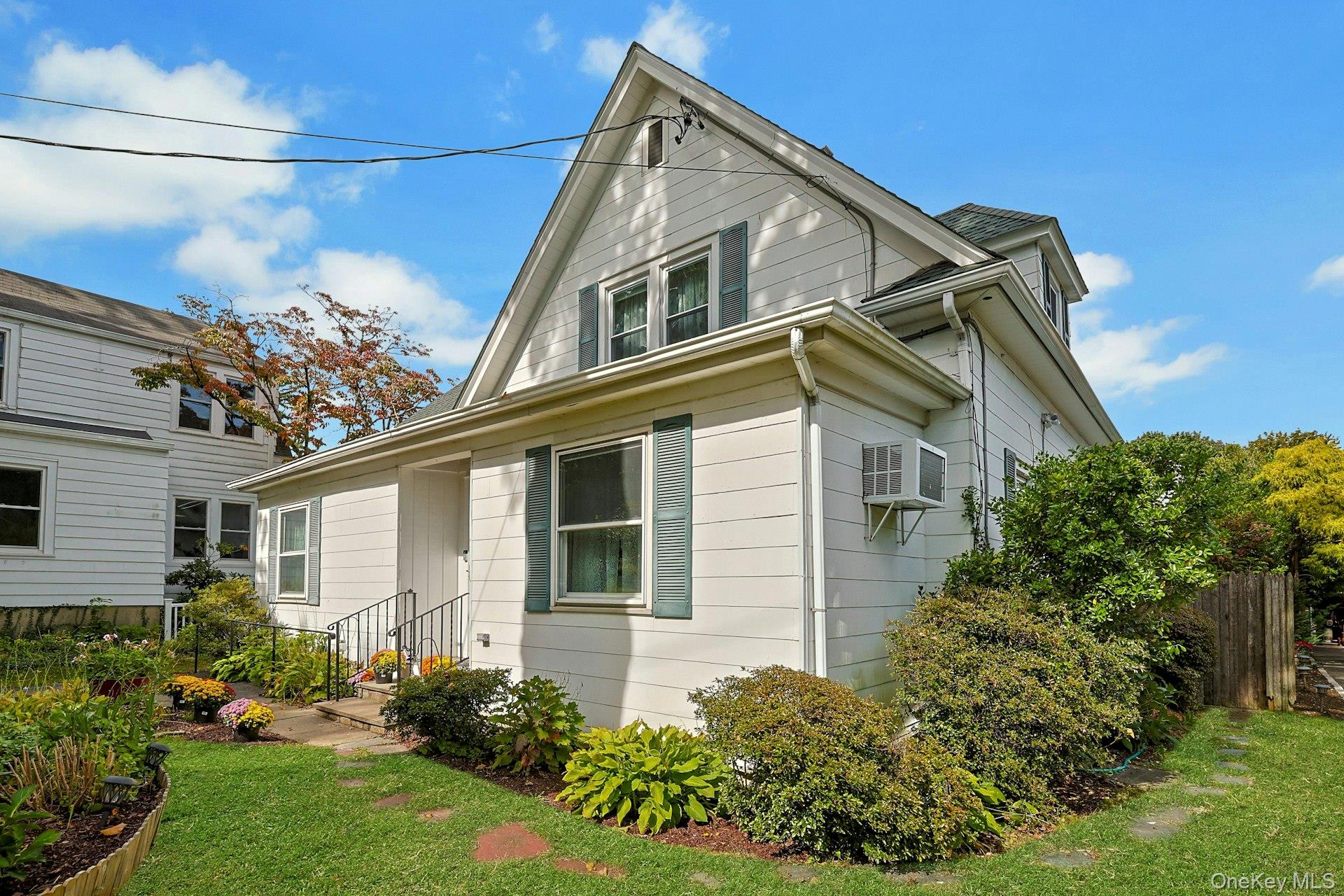 White two-story house with green shutters, flower garden, and neighboring home at left.