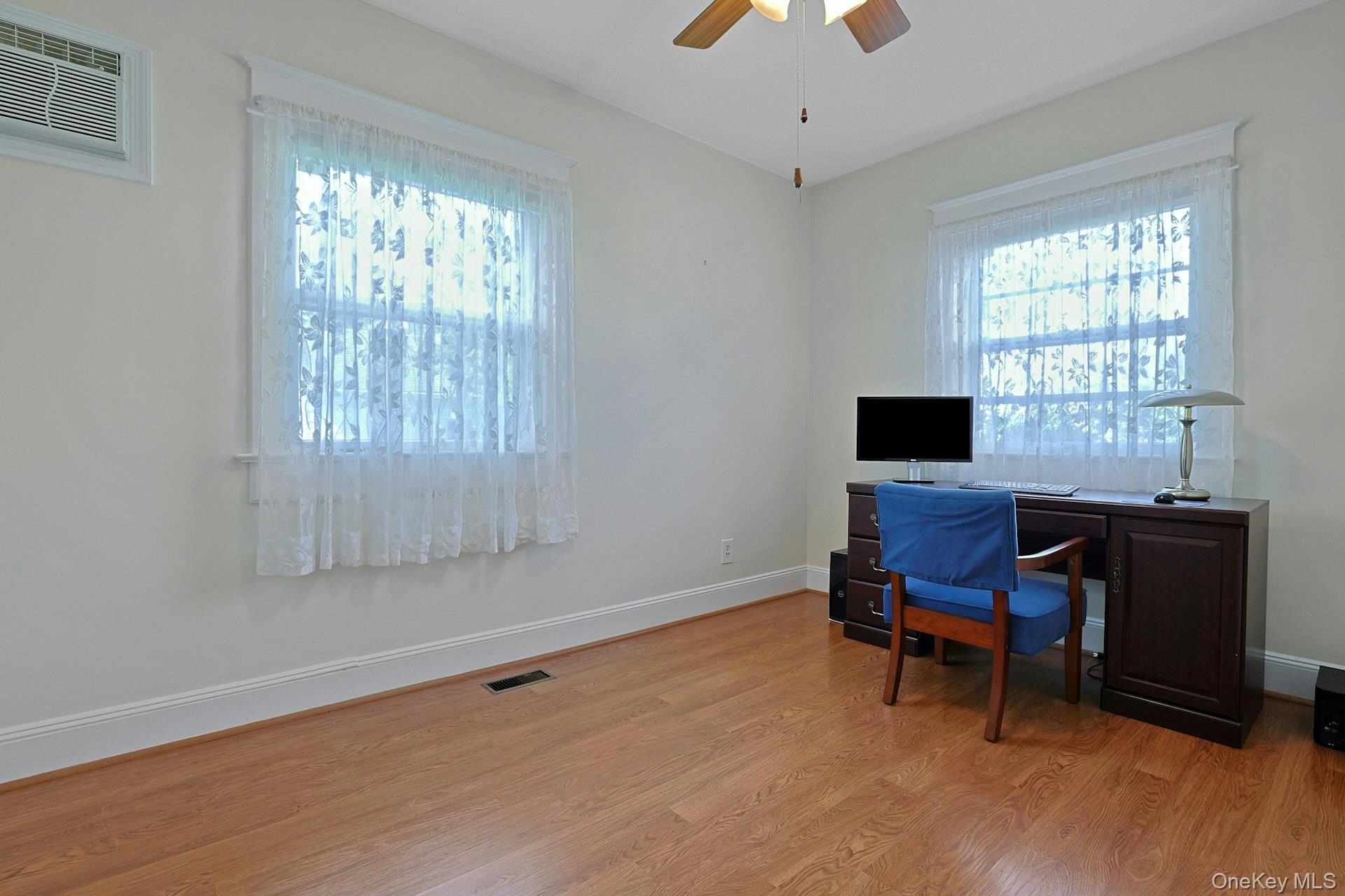 Furnished home office with a dark wood desk, blue chair, and two lace-curtained windows.