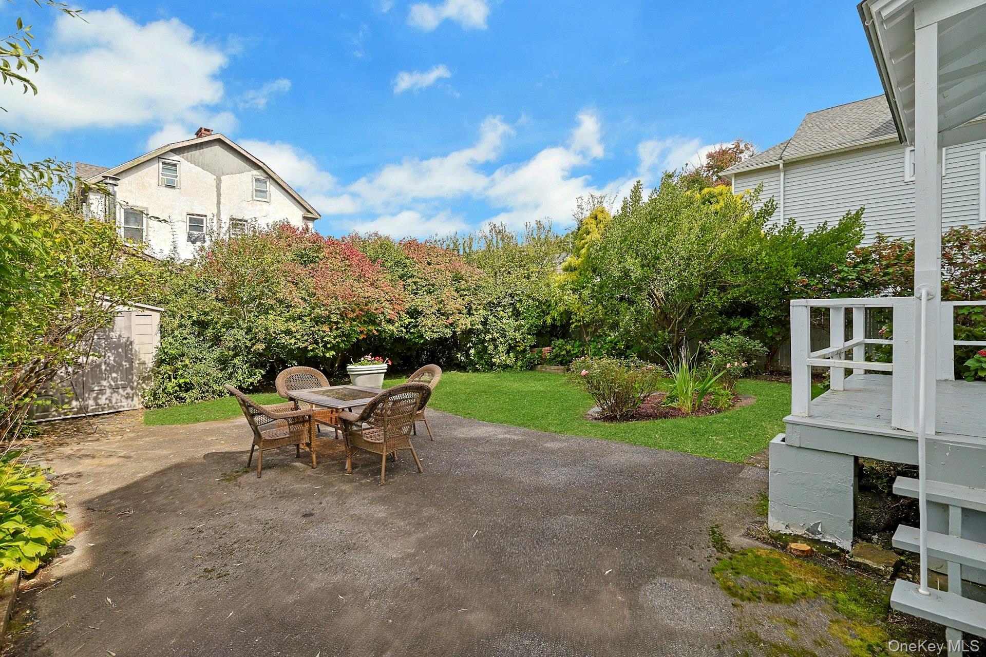 Backyard patio with wicker dining set, green lawn, dense hedges, and a white deck.