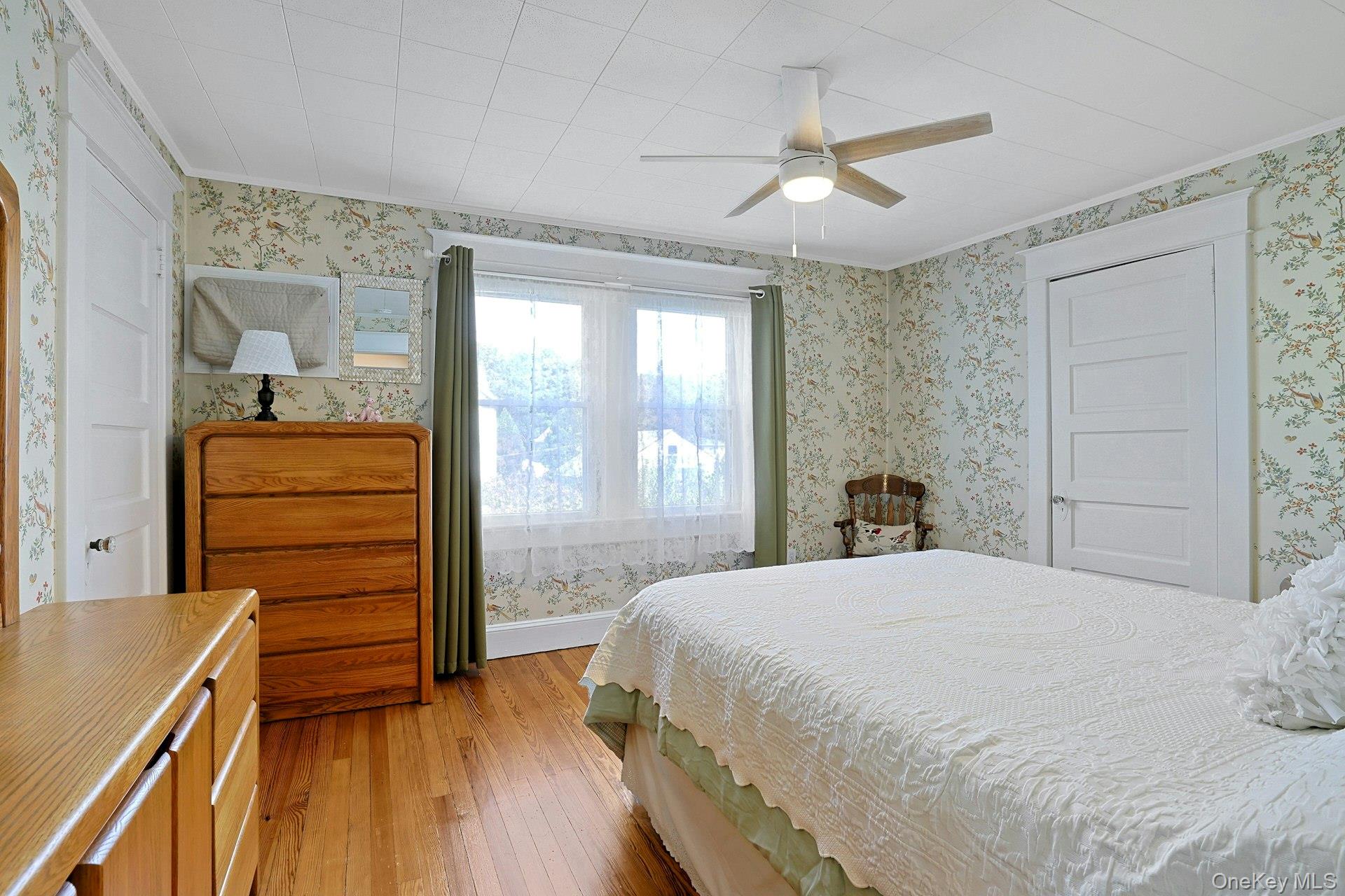 Bedroom with floral wallpaper, hardwood floors, a white bed, and a wooden dresser.
