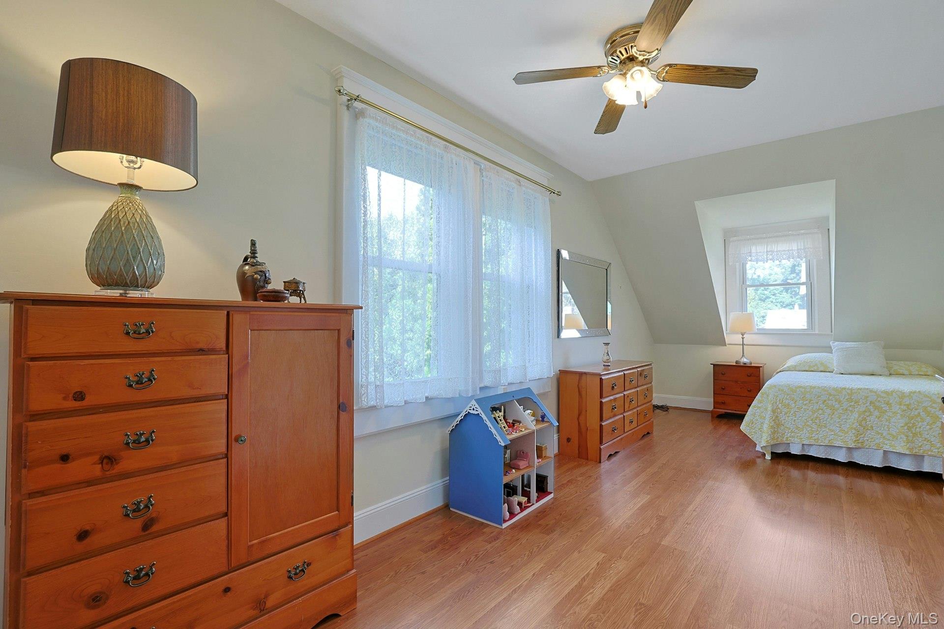 Furnished bedroom with hardwood floors, lace-curtained windows, wooden dressers, and a ceiling fan.
