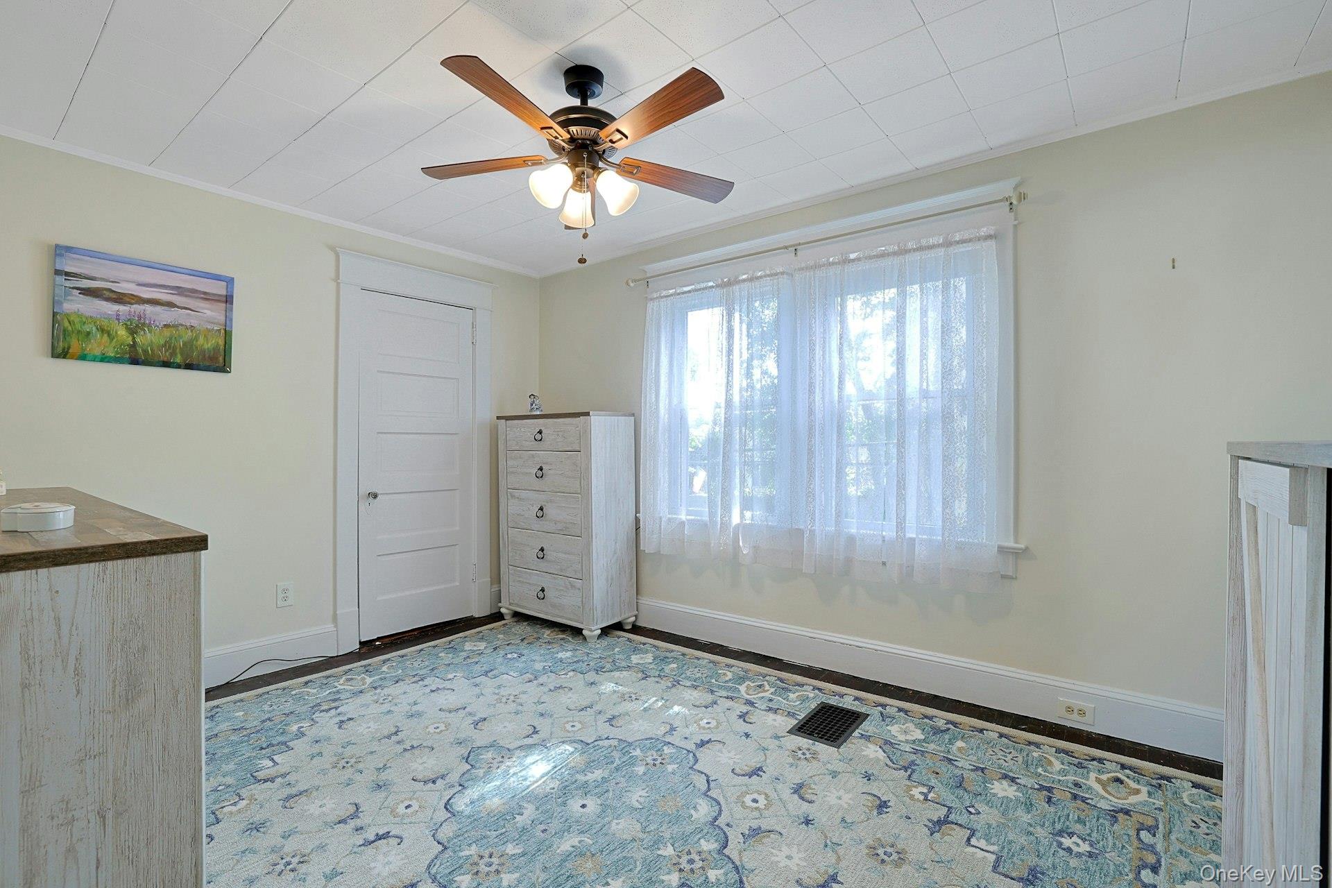 Furnished room with a blue patterned rug, white dresser, and ceiling fan beside a bright window.