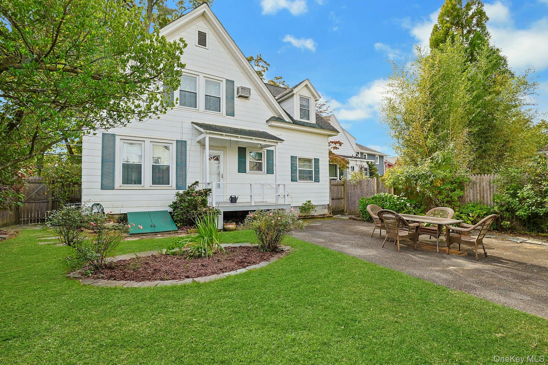 White gabled house with teal shutters, a covered porch, and a patio dining set in a leafy yard.