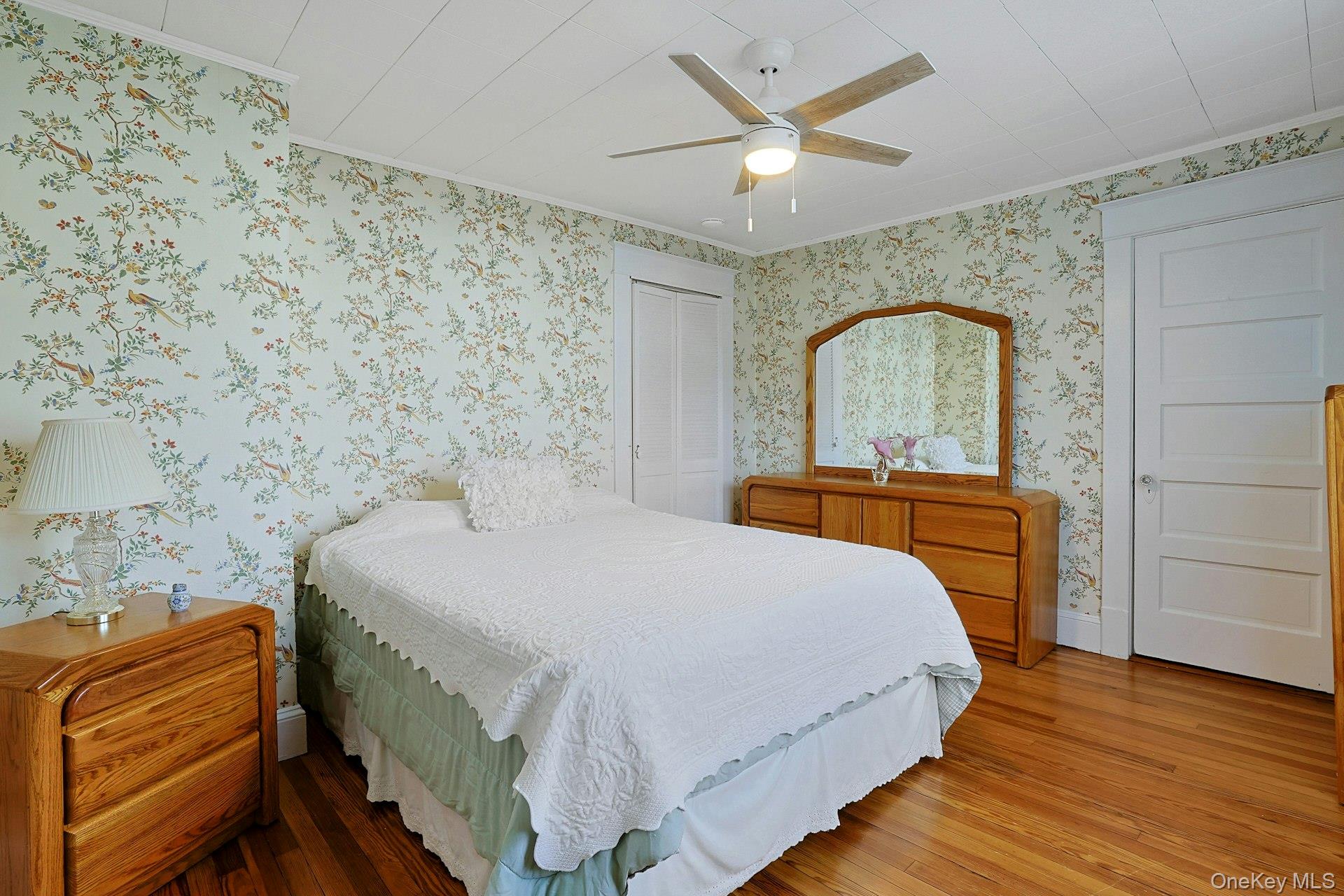 Bedroom with floral wallpaper, hardwood floors, and wooden dressers surrounding a white bed.