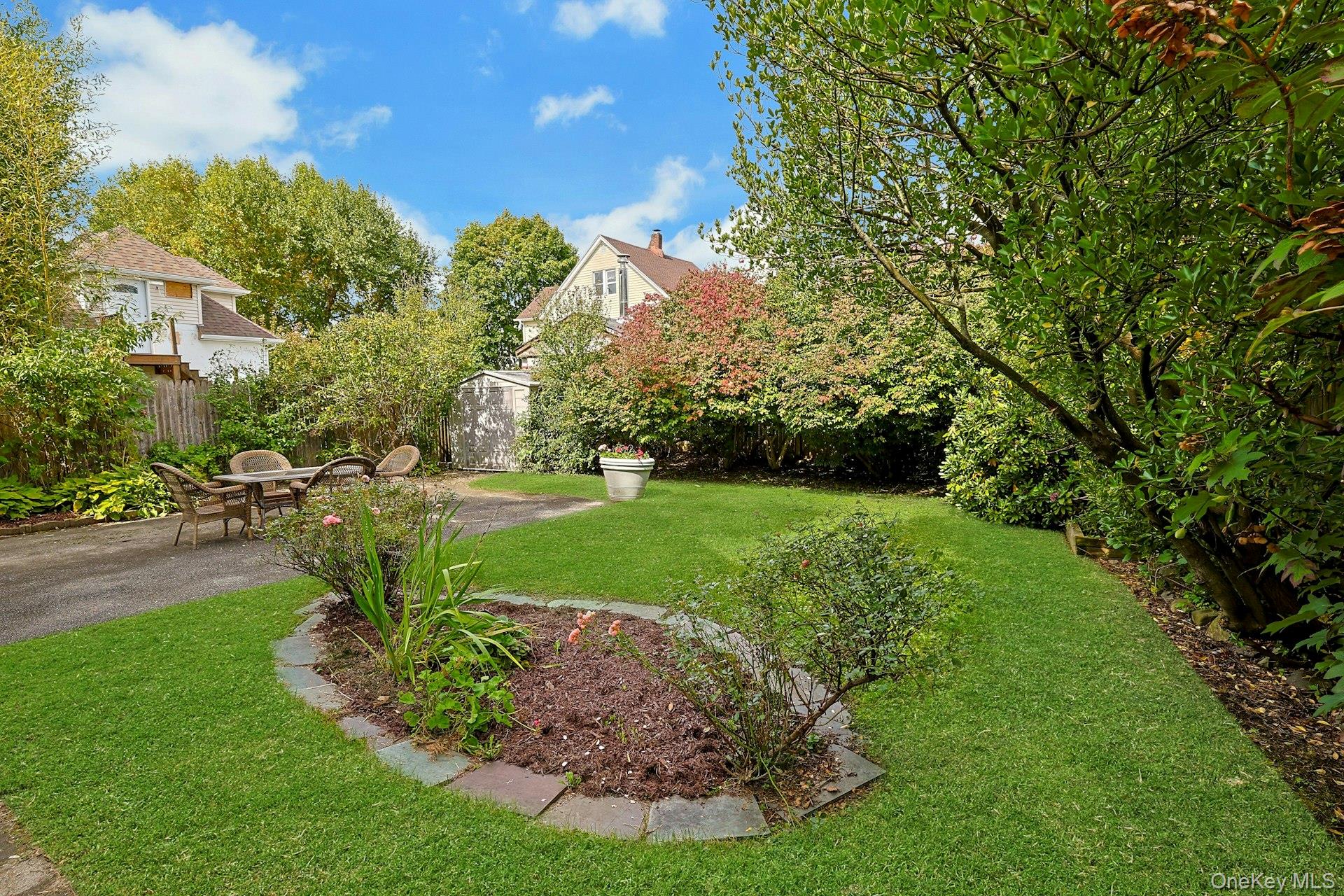 Landscaped backyard with a green lawn, mature trees, garden beds, and a patio seating area.
