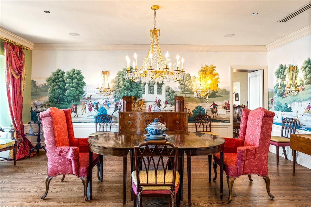 Dining room with an oval table, red chairs, ornate chandeliers, and a garden mural.