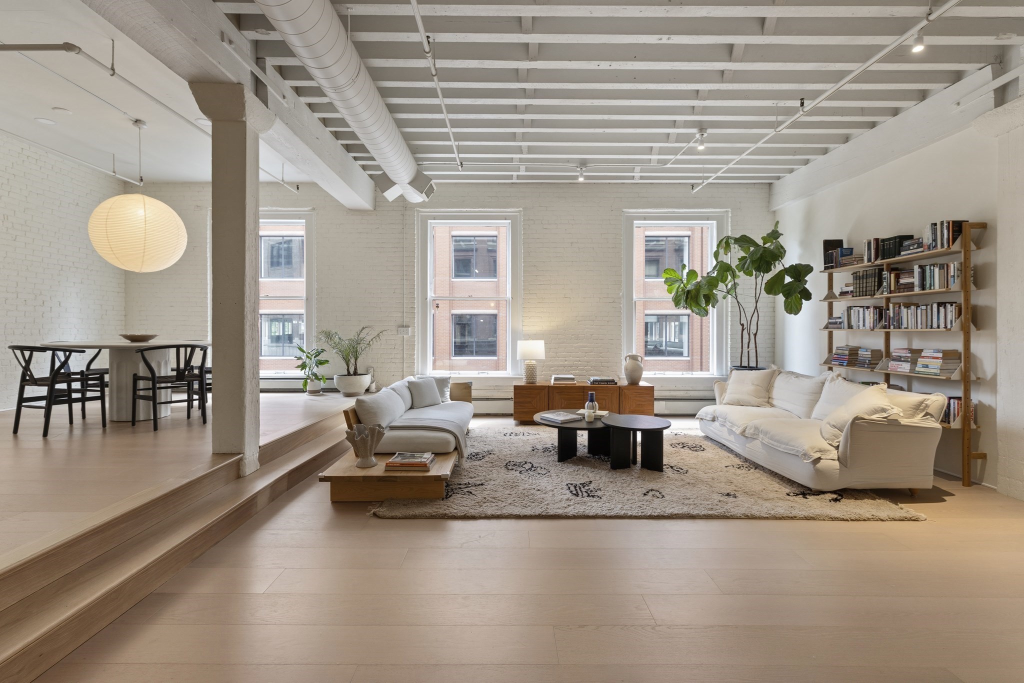 Open-plan loft living room with exposed beams, tall windows, white sofas, and a dining area.