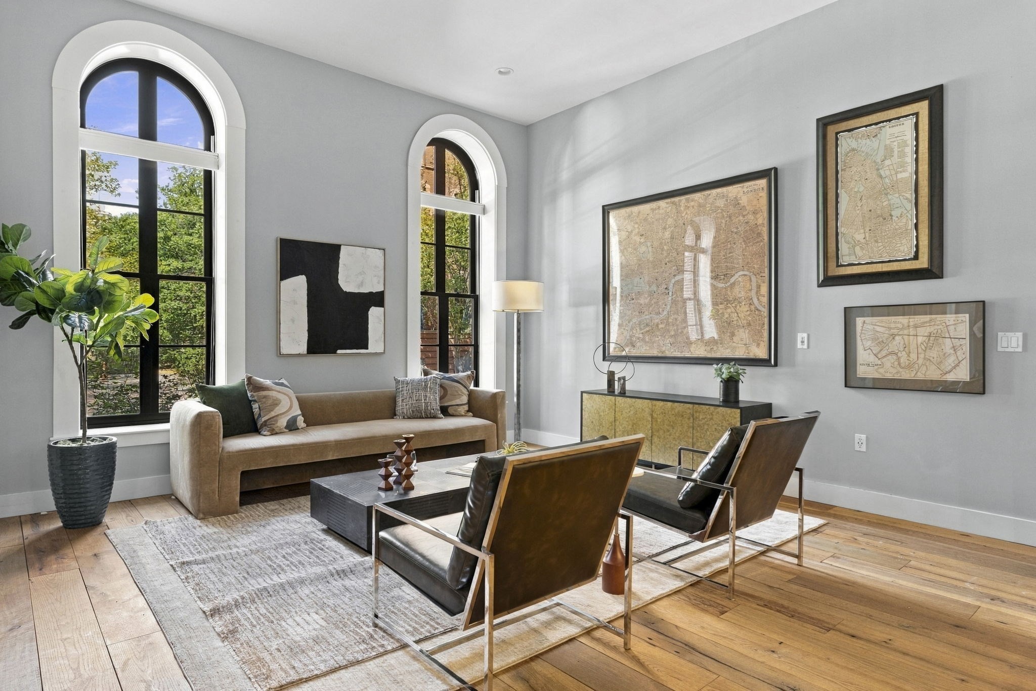 Furnished living room with arched windows, a tan sofa, leather chairs, and framed maps.