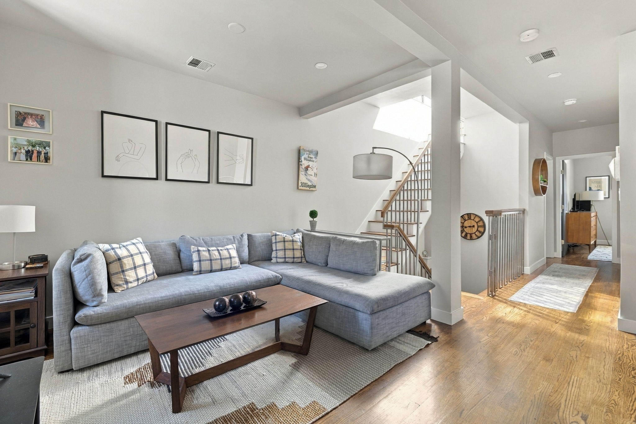 Living room with a gray sectional, plaid cushions, hardwood floors, and a staircase beneath a skylight.