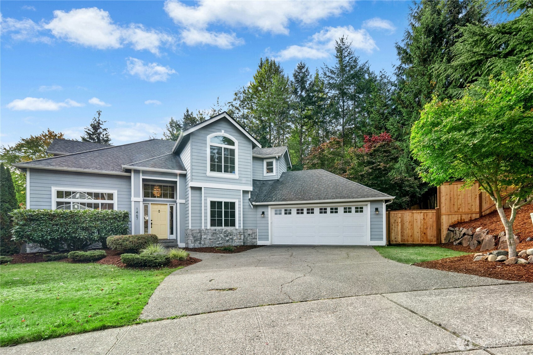 Two-story blue-gray house with a double garage, yellow front door, landscaped lawn, and wooden fence.