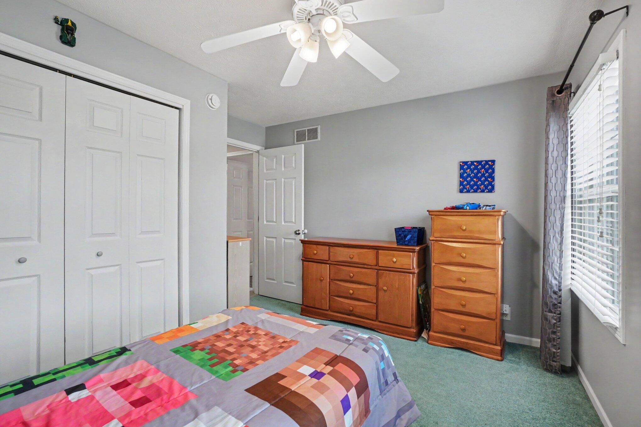 Bedroom with colorful patchwork bedding, wooden dressers, and a window with blinds.