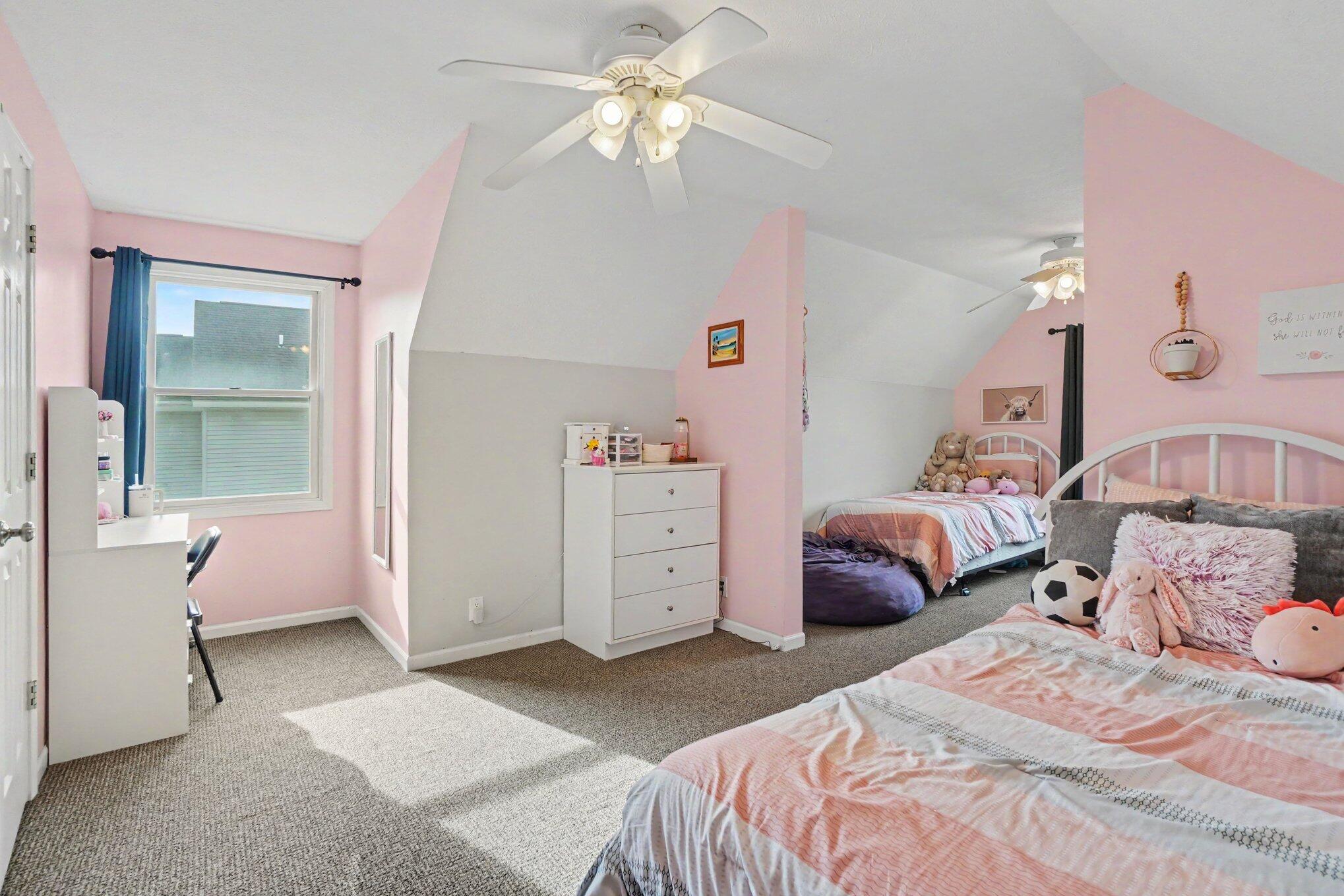 Furnished attic bedroom with pink walls, two beds, and a desk beside the window.