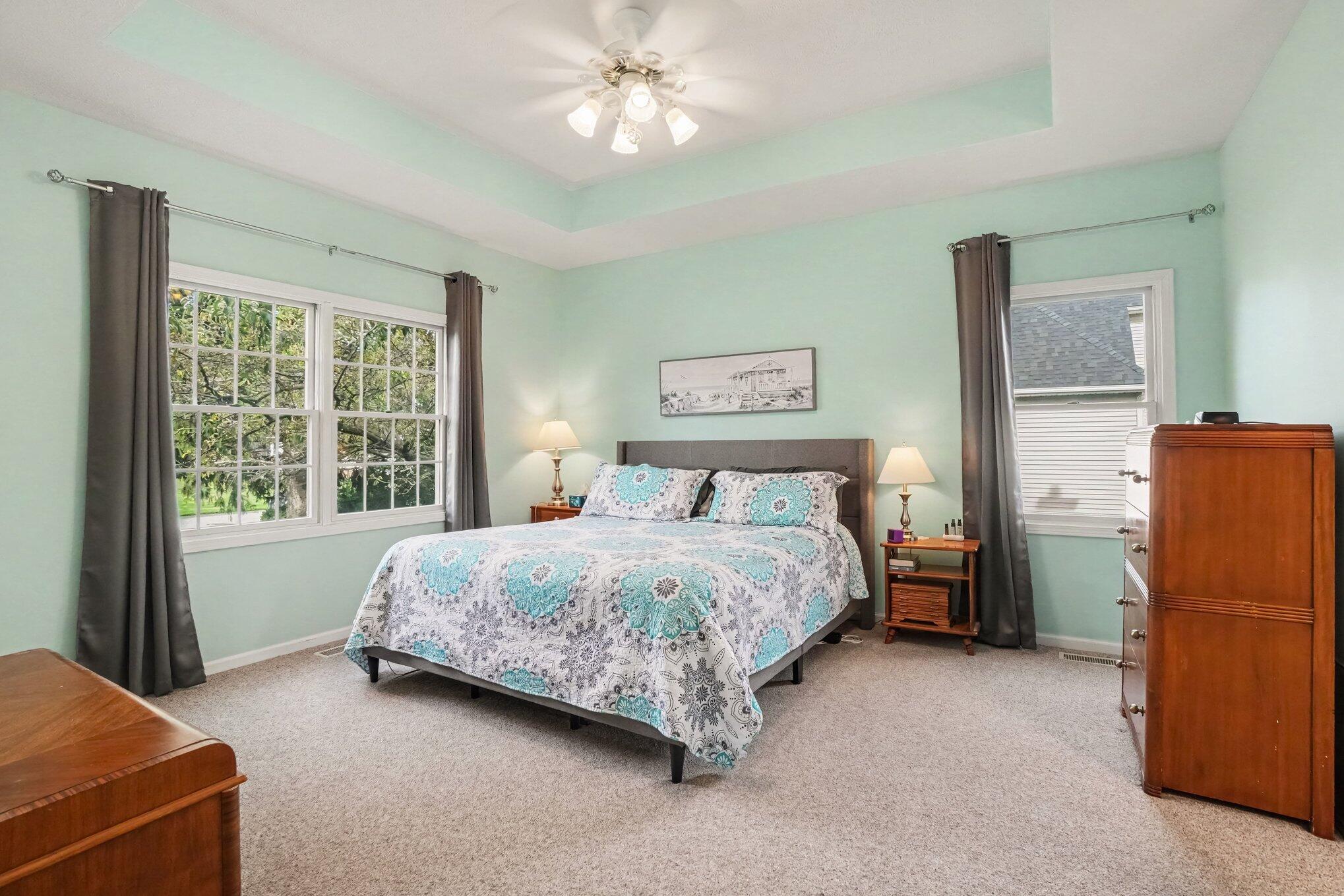Bedroom with turquoise-patterned bedding, a tray ceiling, and windows with dark curtains.