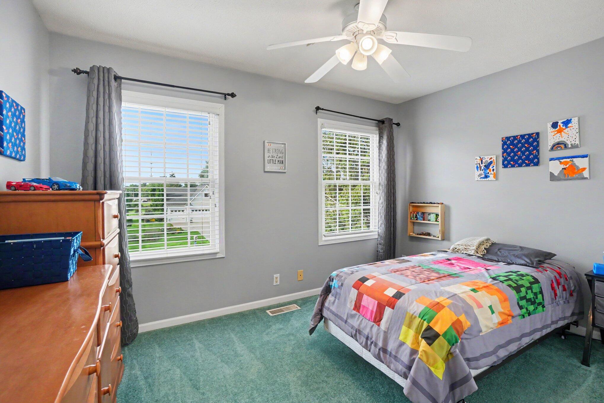 Bedroom with teal carpet, two shuttered windows, and a bed covered in a colorful patchwork quilt.