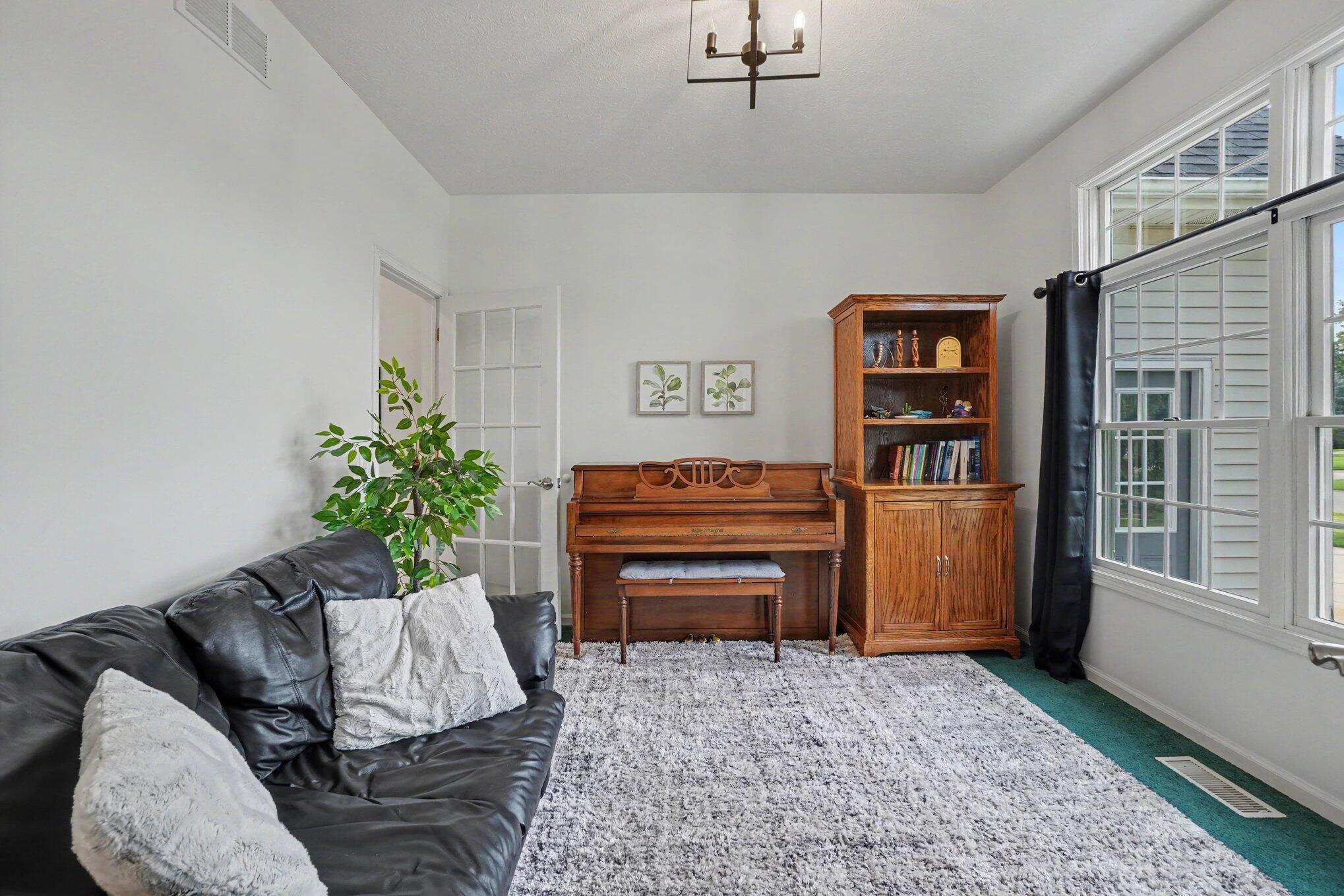 Living room with an upright piano, wooden bookcase, black leather sofa, and large windows.