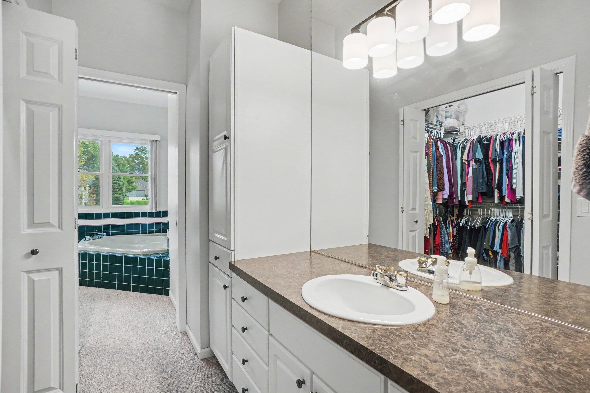 Bathroom with double-sink vanity, teal-tiled soaking tub, and open clothing closet.