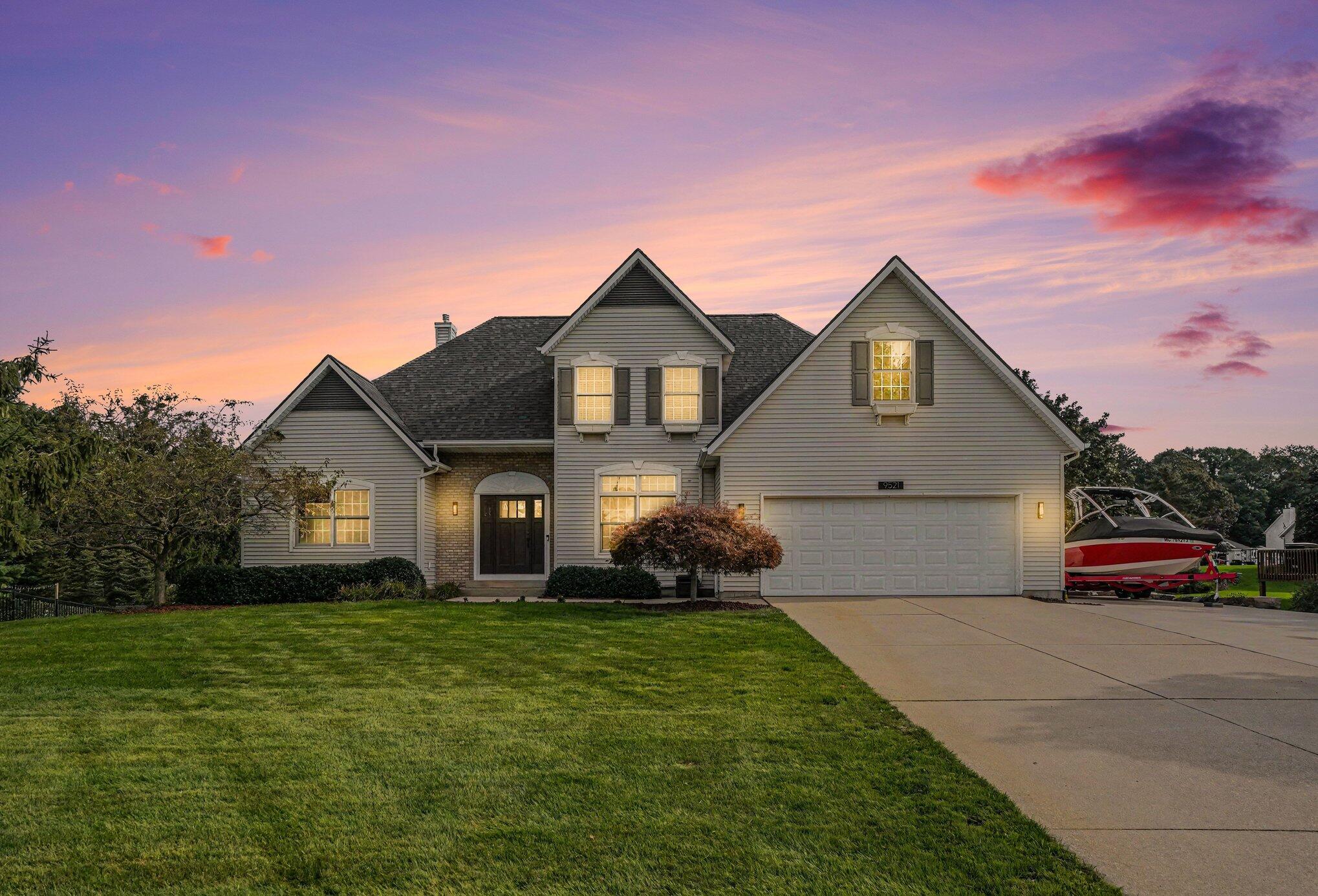 Gray two-story home with a double garage and a red-and-white boat parked beside it at sunset.