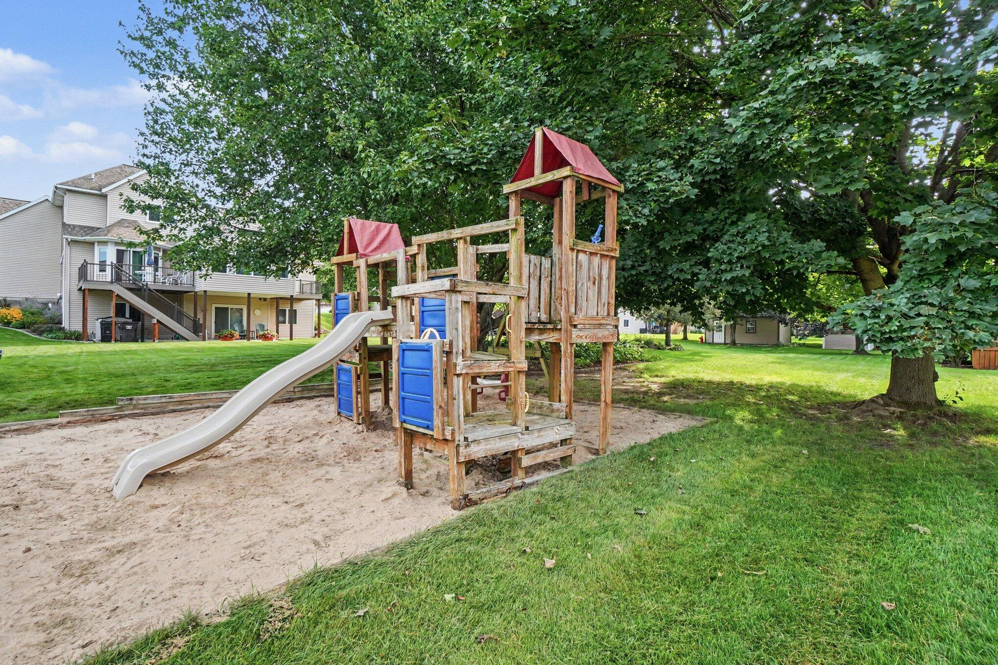 Backyard play structure with a slide over a sand play area, surrounded by mature trees and lawn.