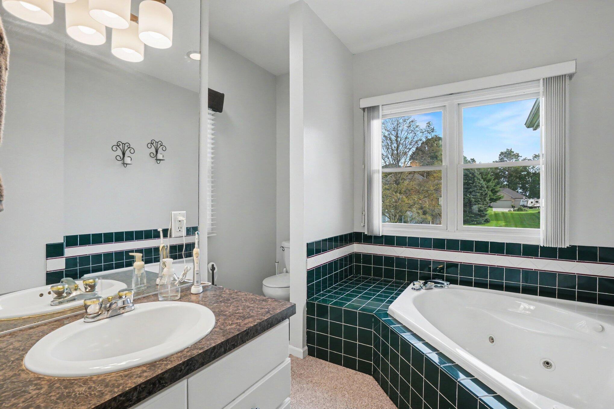 Bathroom with a jetted tub in a dark green tile surround and a wide vanity beneath a large mirror.