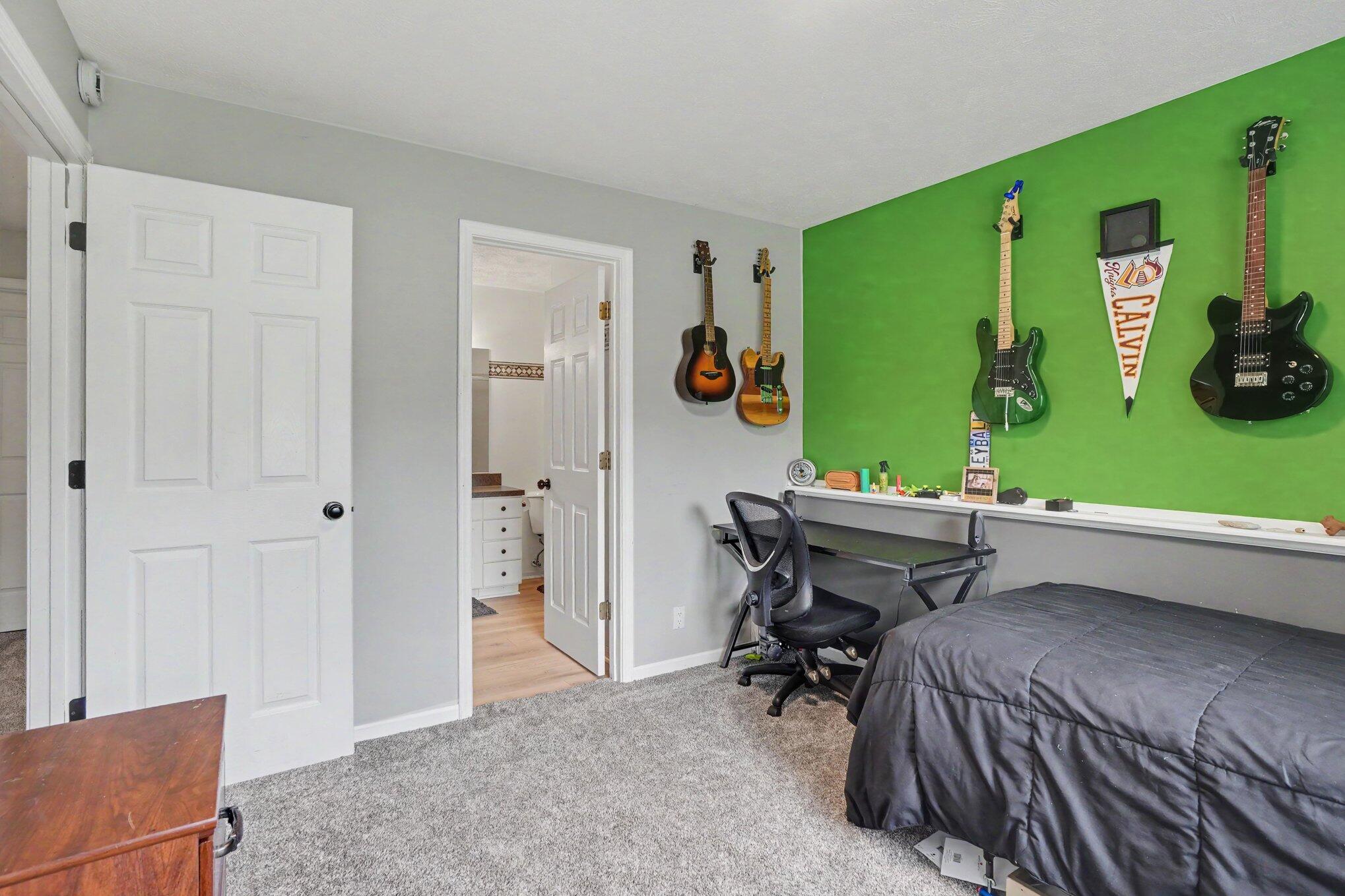Bedroom with a bright green guitar display wall, desk, and gray-carpeted floor.