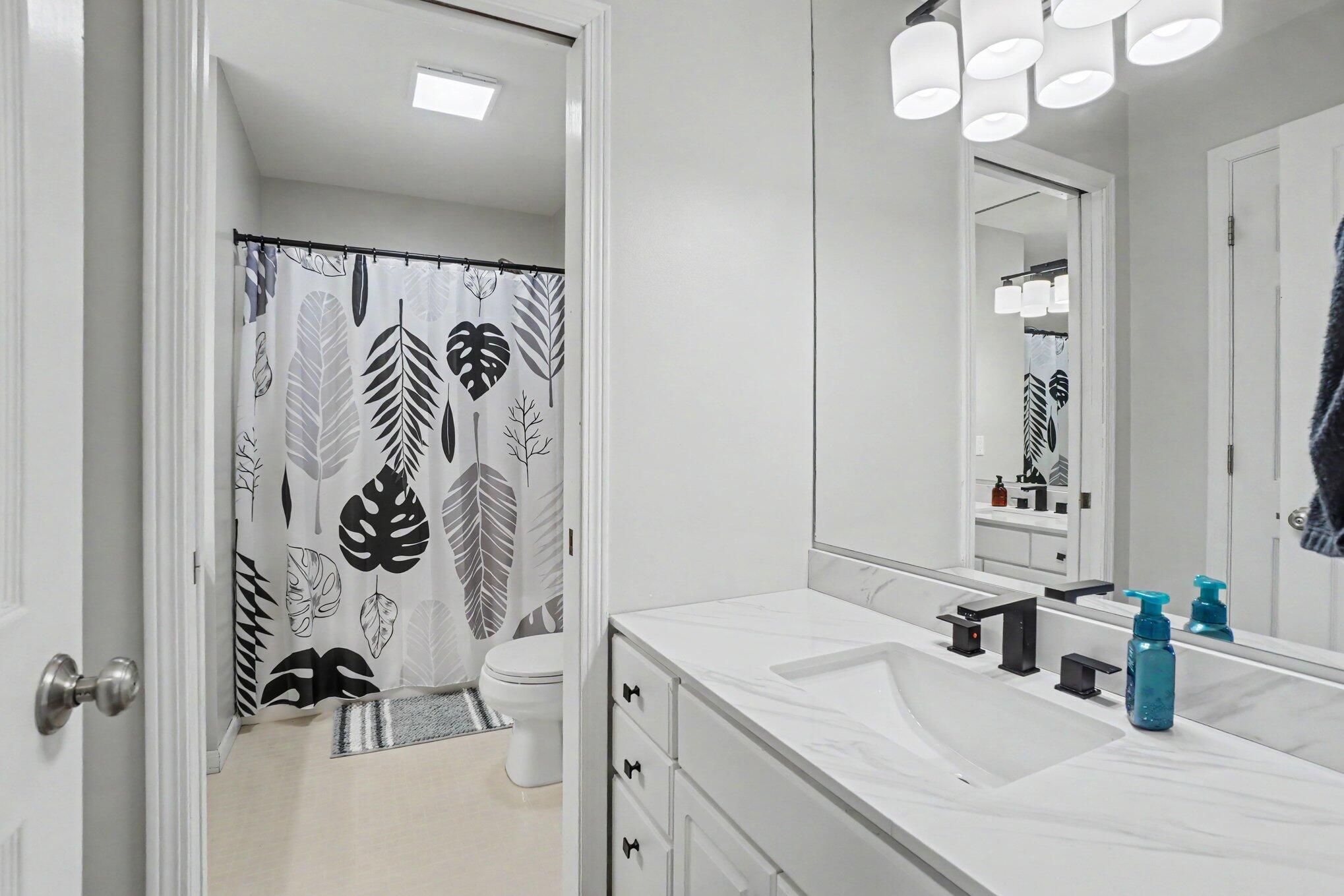 Bathroom with a white marble-look vanity and mirror beside a leafy-patterned shower curtain.