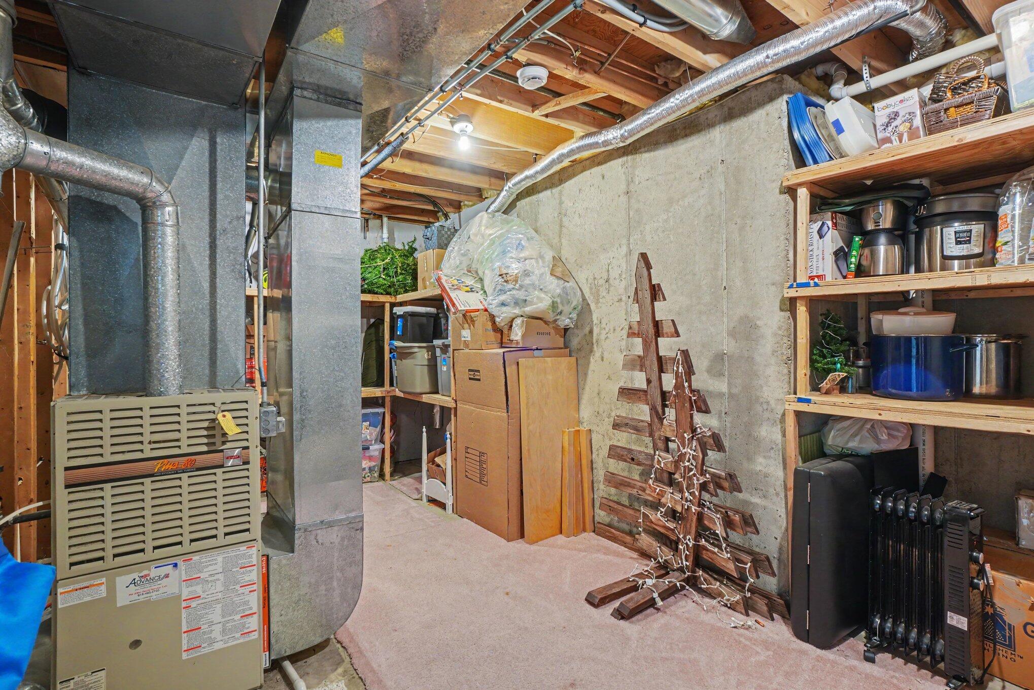 Basement utility area with a large furnace, exposed ductwork, concrete walls, and storage shelves.