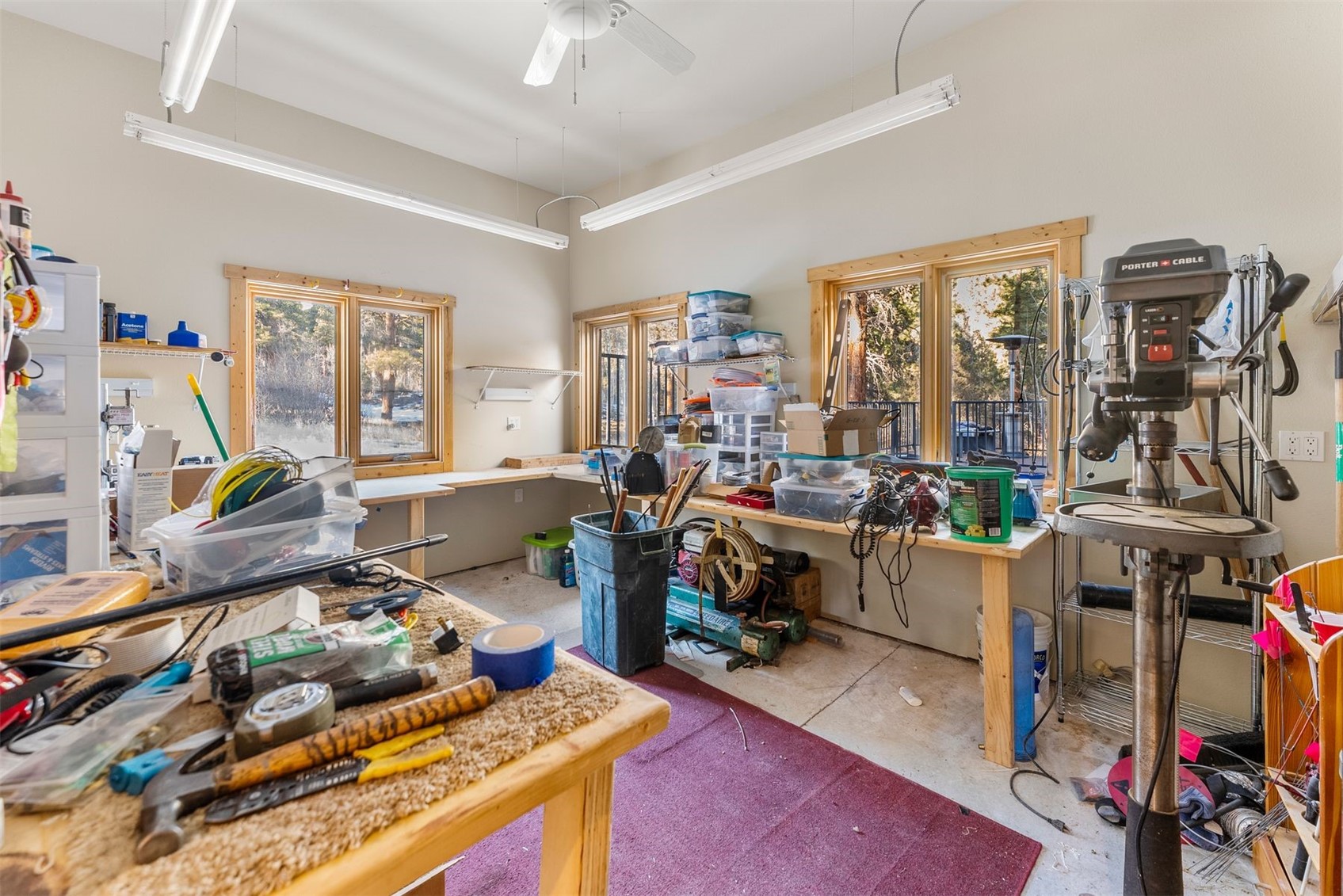Workshop with crowded workbenches, storage bins, and a drill press beside wood-trimmed windows.