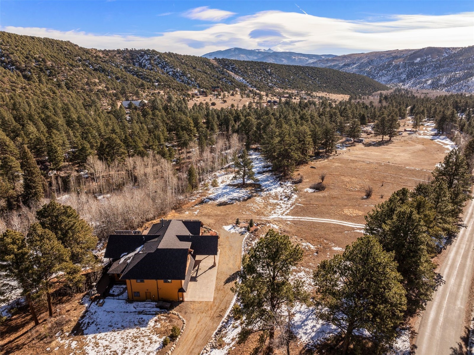 Aerial view of a house beside a snowy meadow, surrounded by pine forest and mountain views.